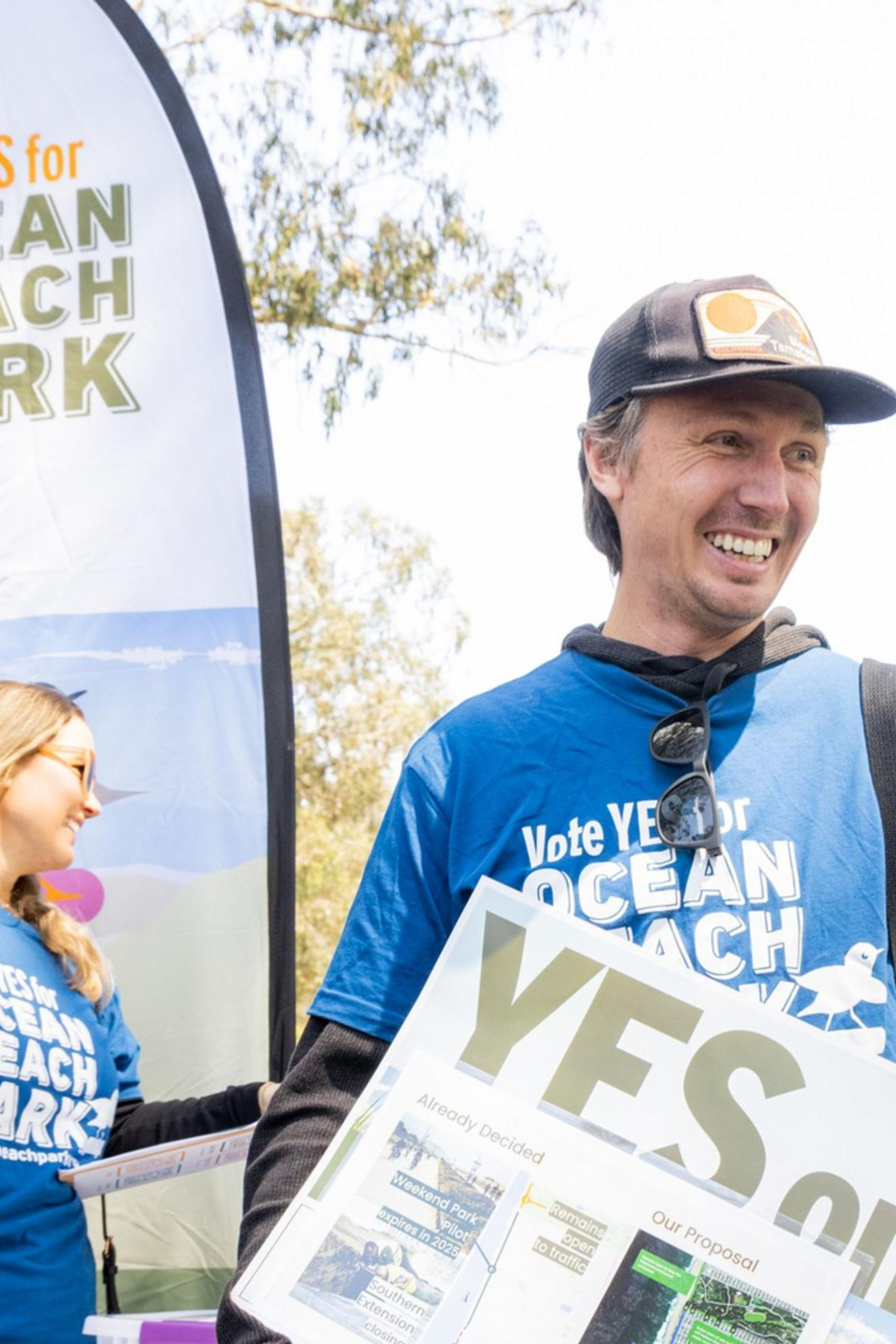 People wearing blue "Vote YES for Ocean Beach Park" shirts are gathered, holding signs supporting Measure K at an outdoor event, with trees and banners in the background.
