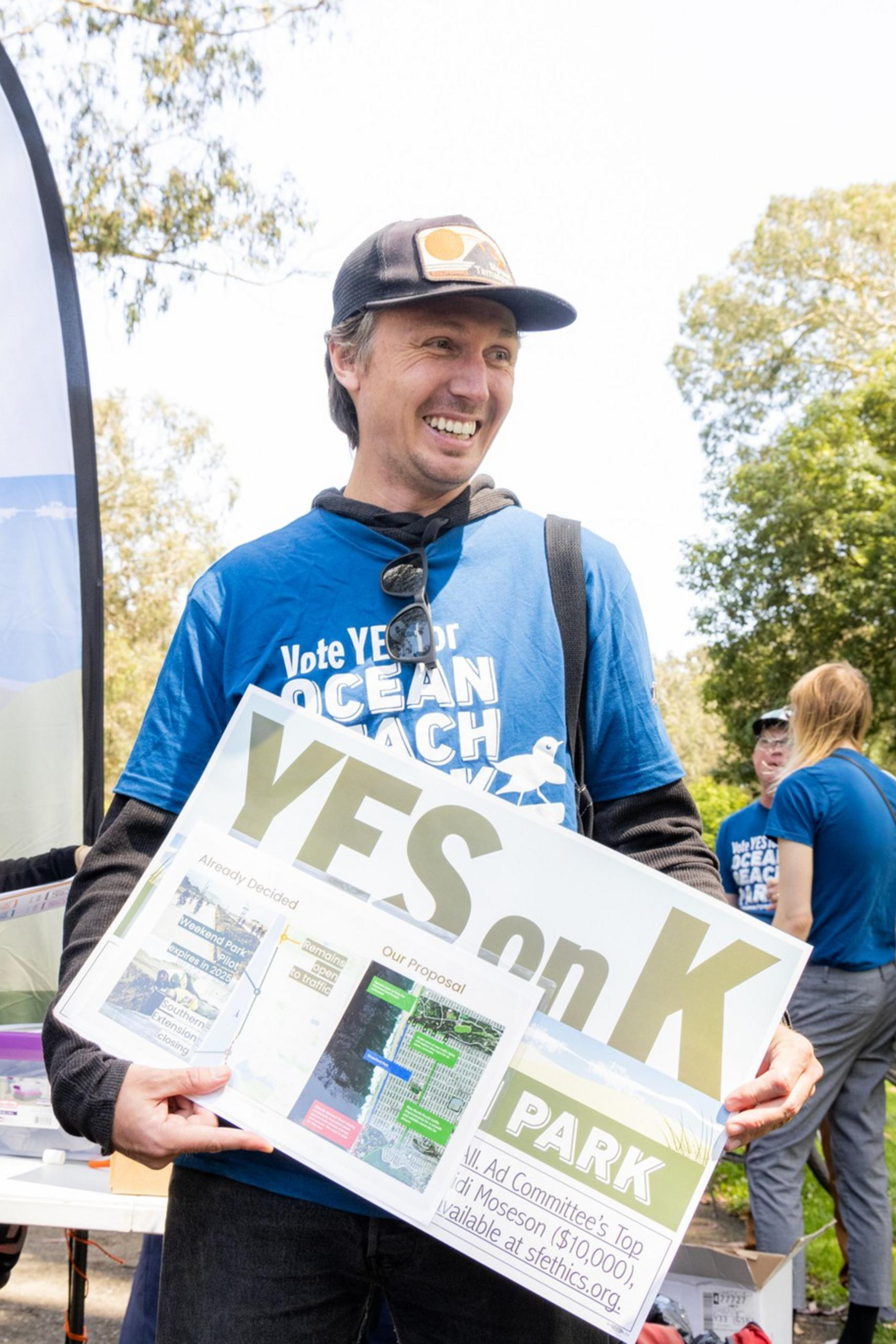 People wearing blue "Vote YES for Ocean Beach Park" shirts are gathered, holding signs supporting Measure K at an outdoor event, with trees and banners in the background.