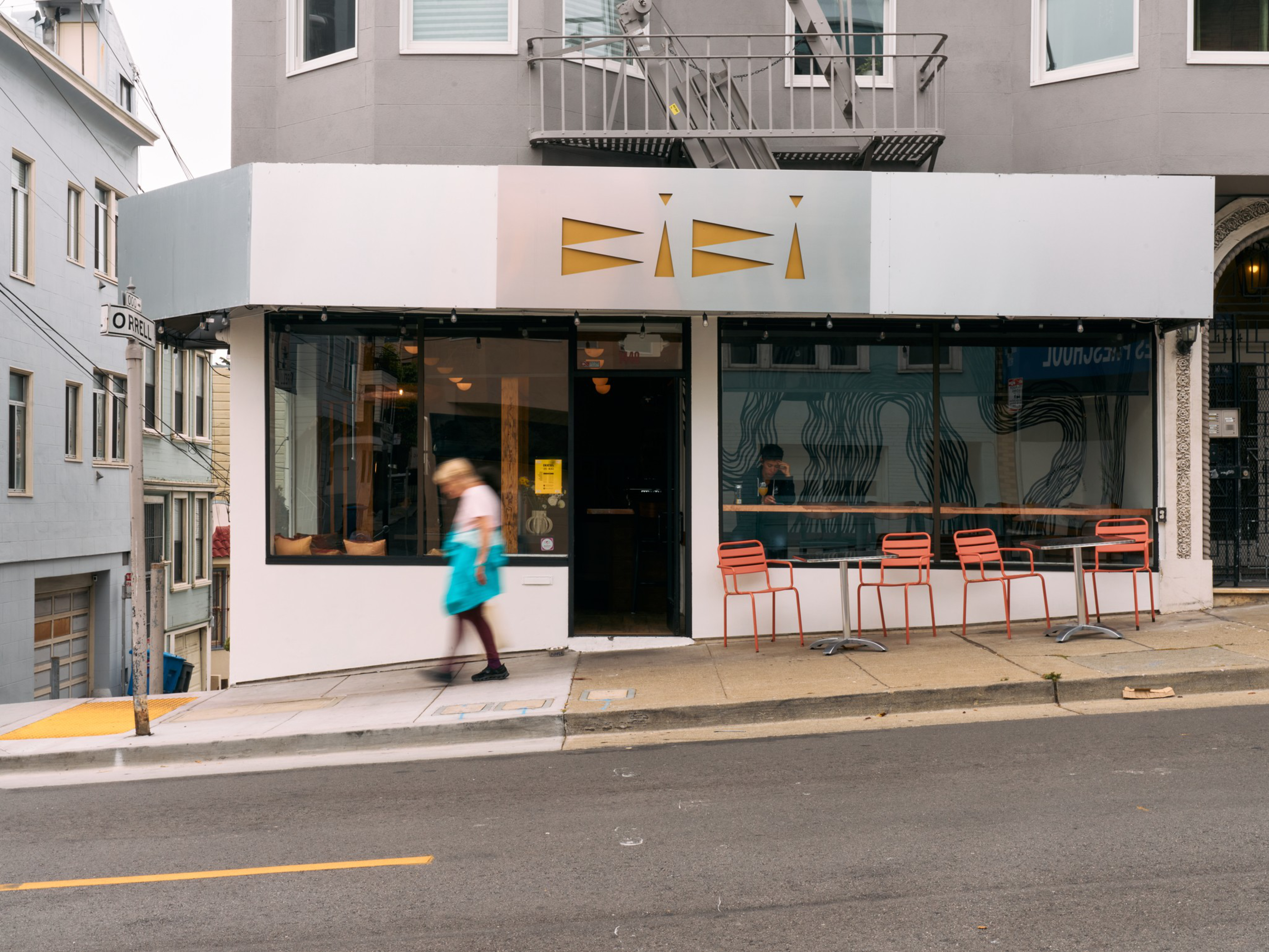 A person in a white shirt and teal apron walks past a small café with orange chairs on a sloped city street.