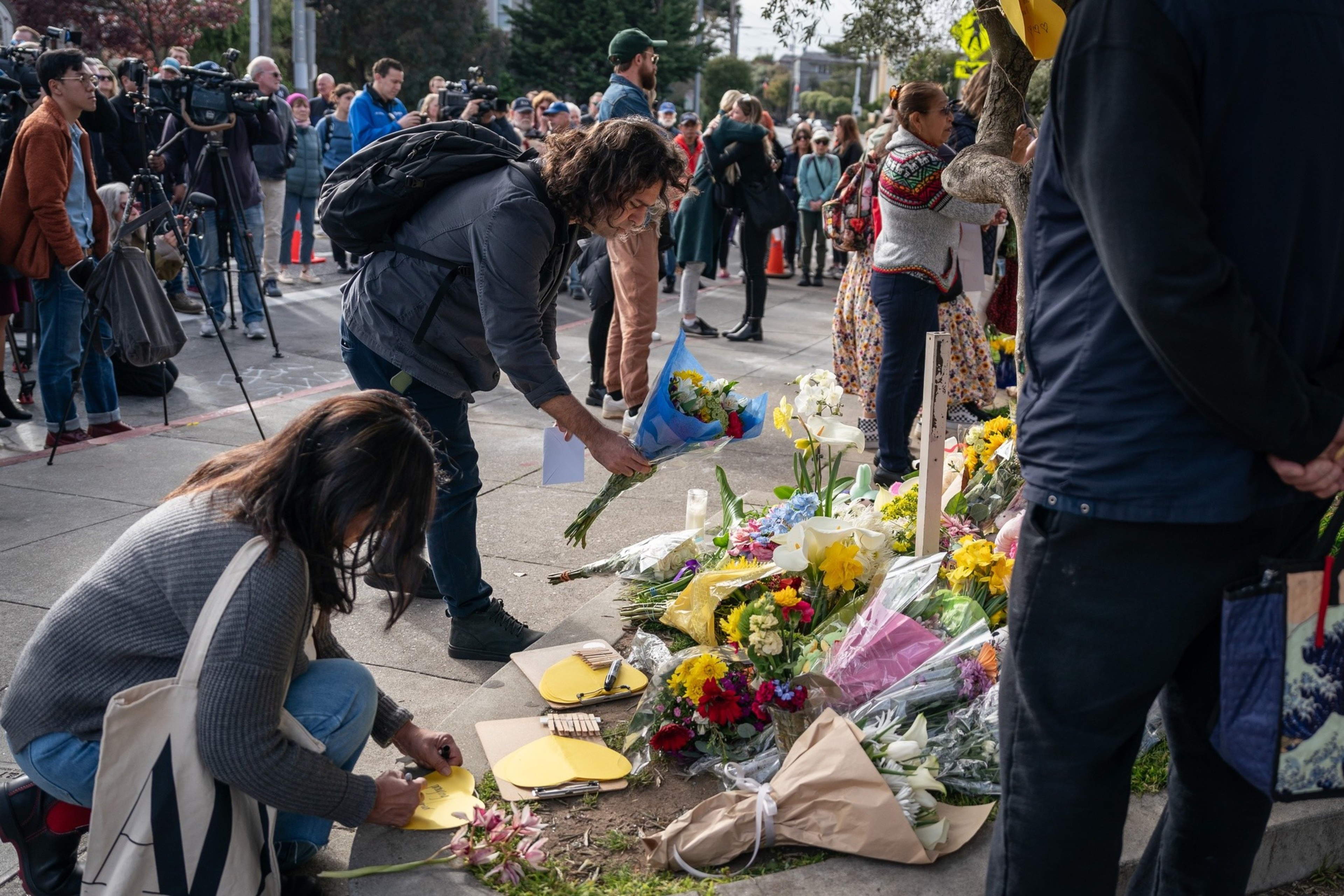 People are placing flowers and notes at a makeshift memorial on a sidewalk. A crowd and media are present.