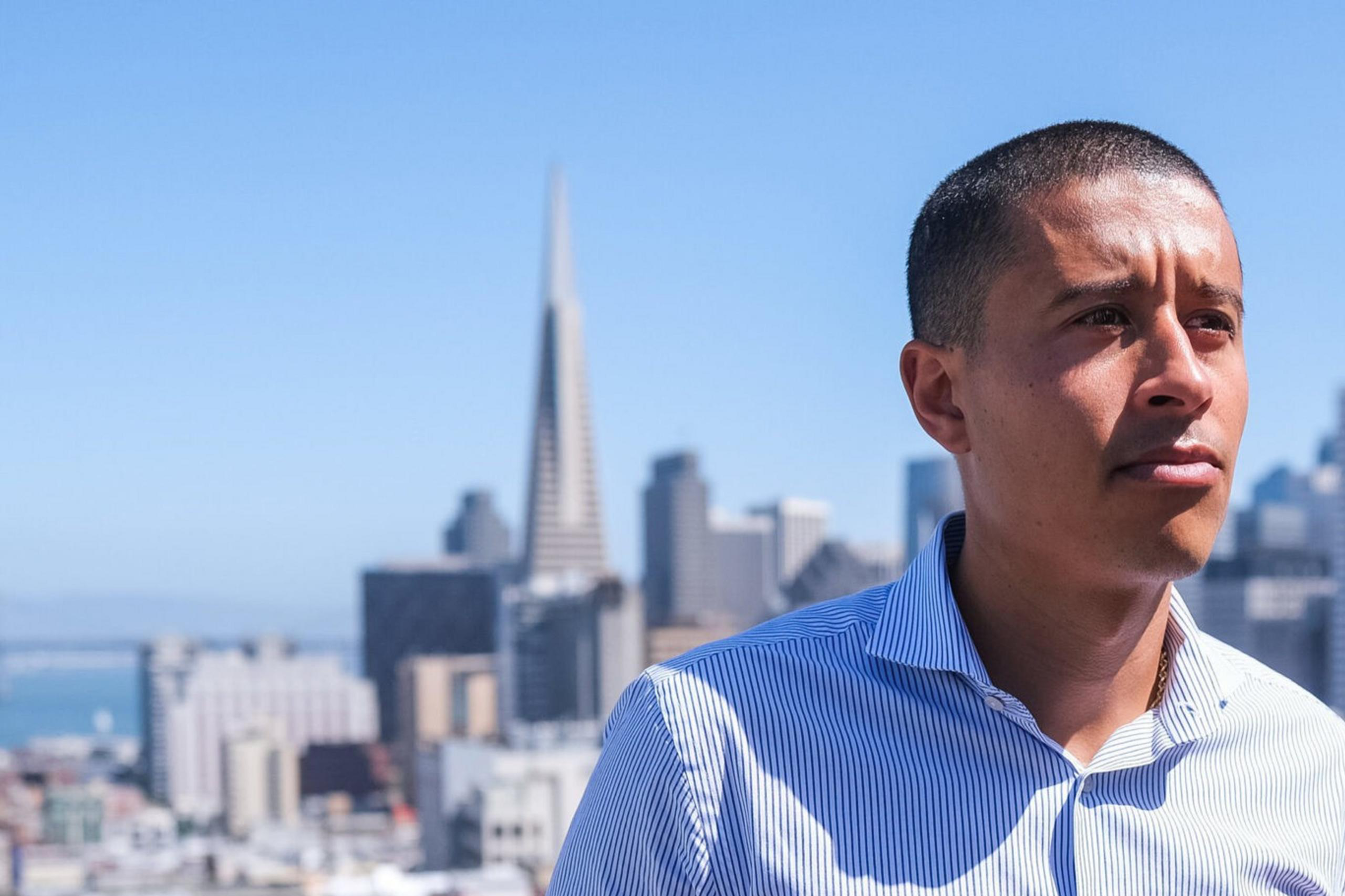 A man in a striped shirt stands on a rooftop with a city skyline in the background, featuring the Transamerica Pyramid under a clear blue sky.