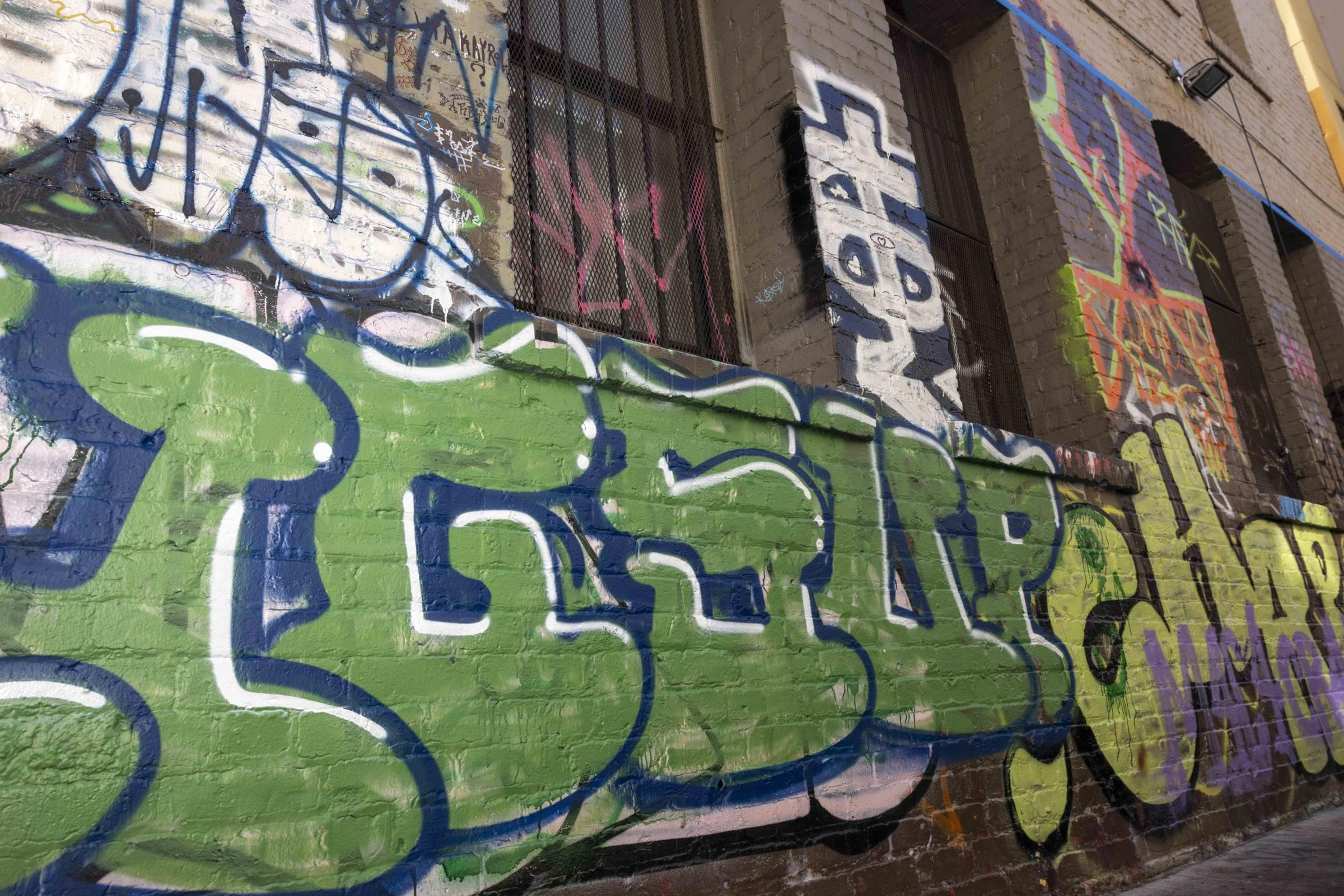 A vibrant graffiti mural covers a brick wall, showcasing bold green and blue letters. Background tags and abstract designs add layers of urban artistry.