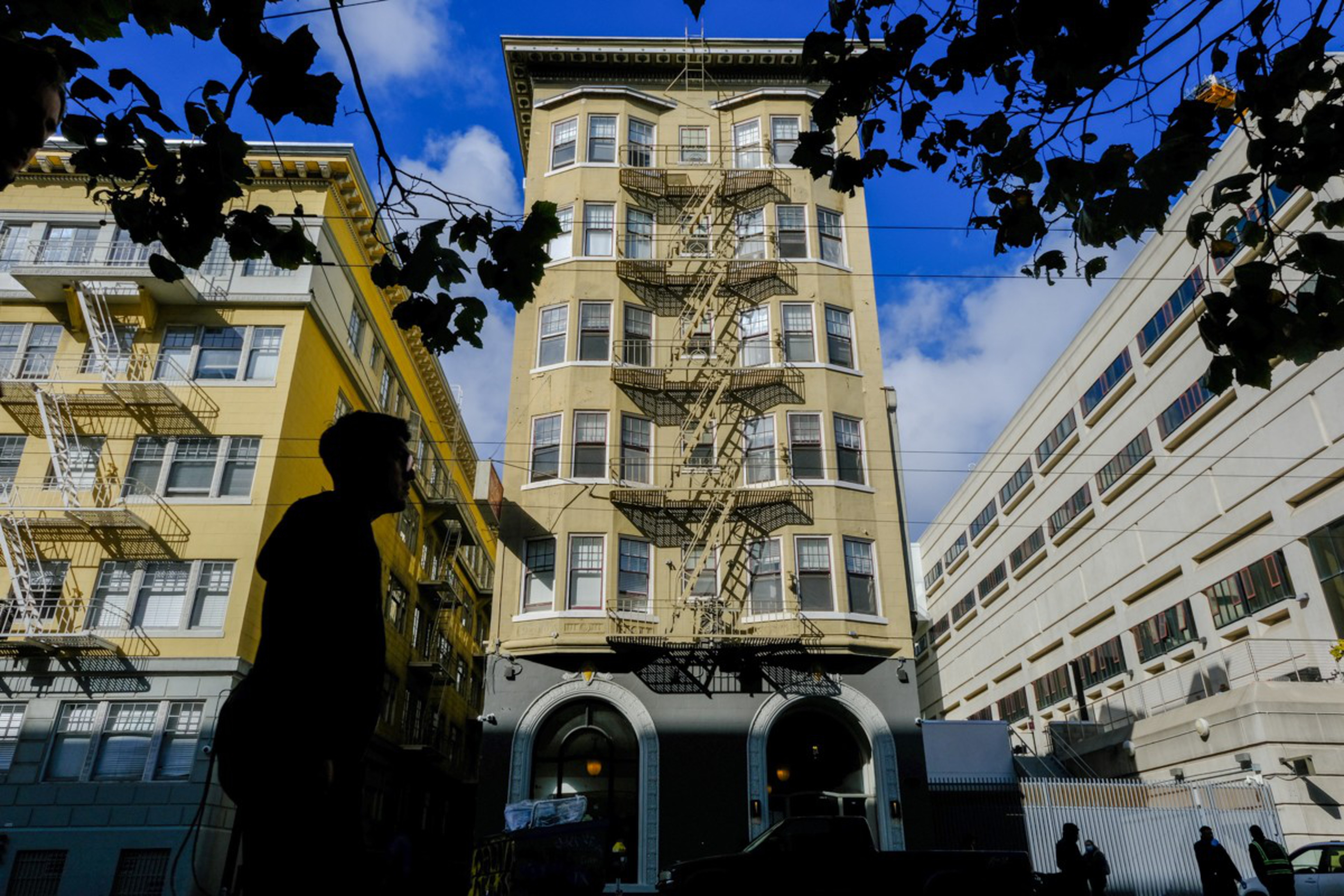 A person’s dark silhouette stands in front of a beige building with fire escapes and a partly cloudy blue sky overhead.