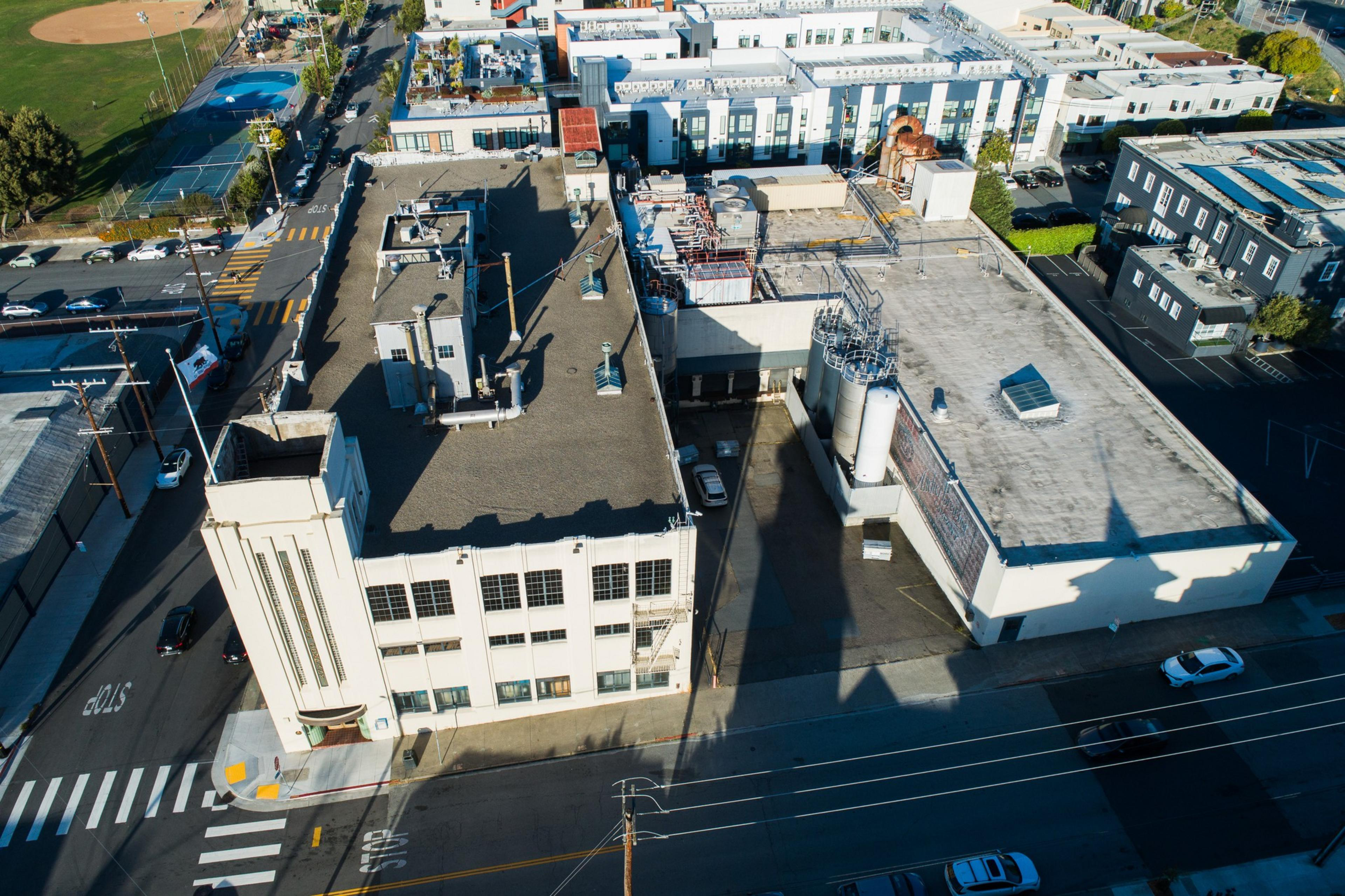 An aerial view shows an industrial building with a flat roof, surrounded by streets. Cars are parked nearby, and a sports field is visible in the background.
