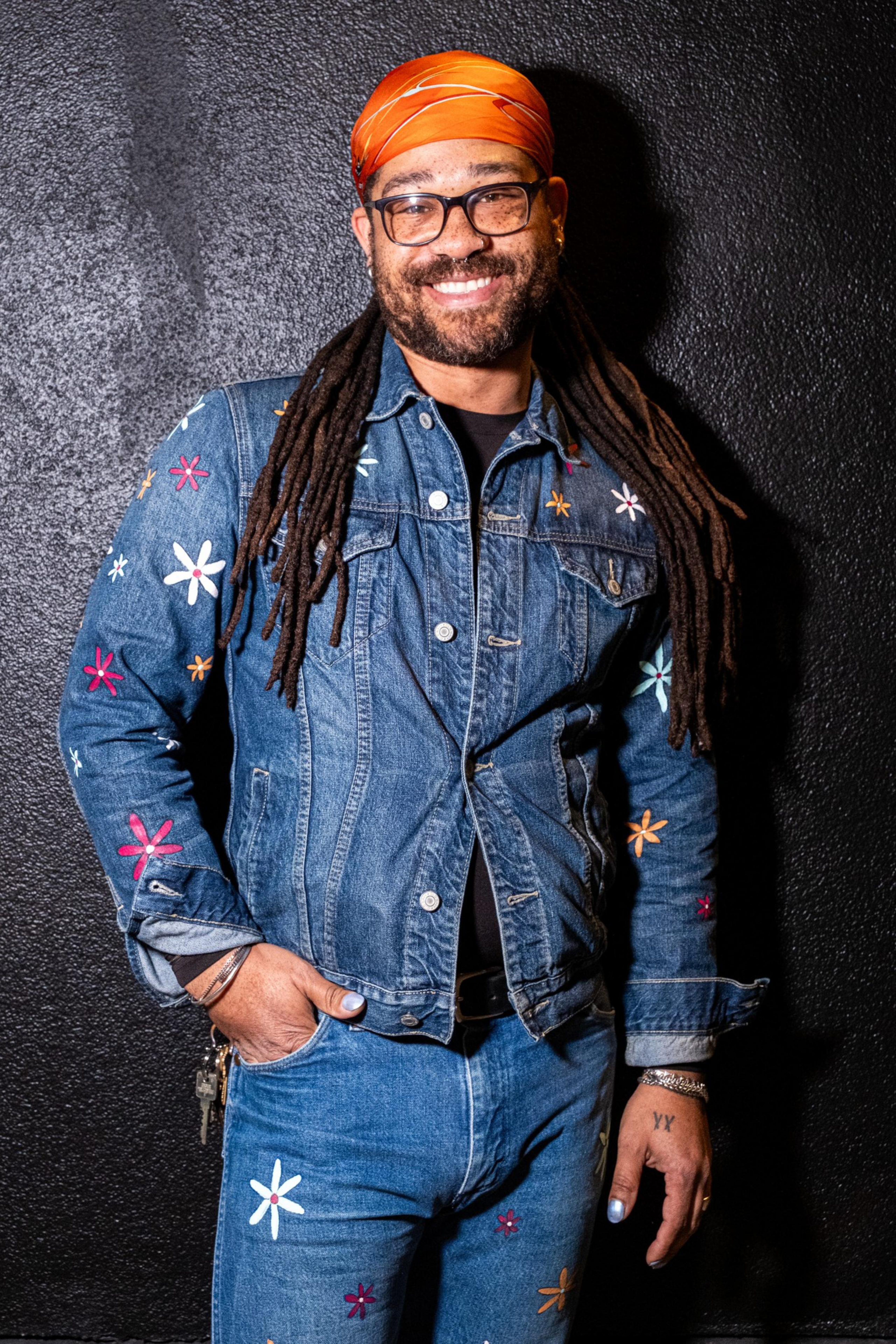 A man smiles wearing a denim jacket and jeans adorned with colorful flower patterns. He also has an orange bandana, glasses, and dreadlocks.