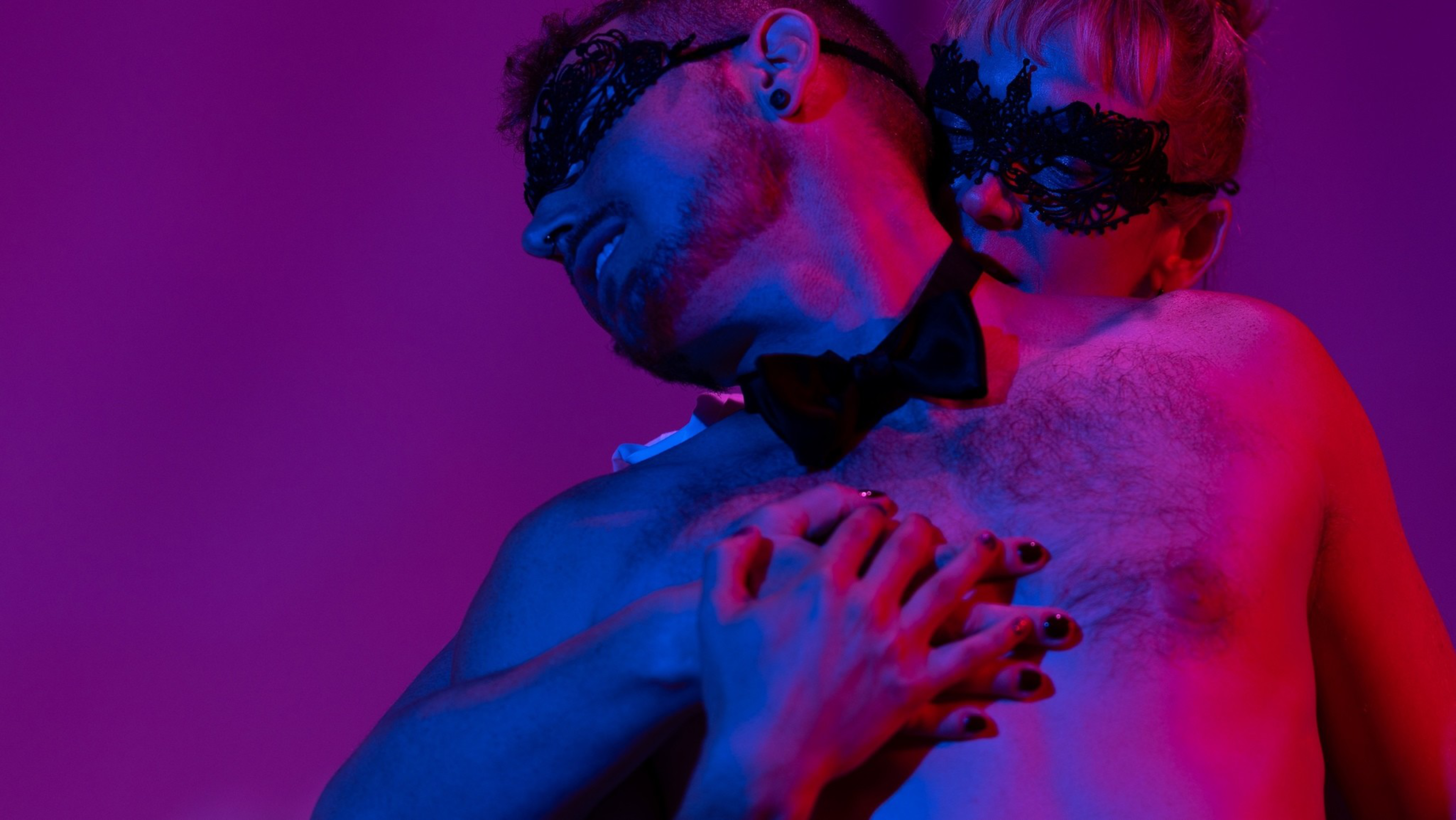 A shirtless man in a bow tie and black lace mask is embraced from behind by a person also wearing a black lace mask, under blue and pink lighting.