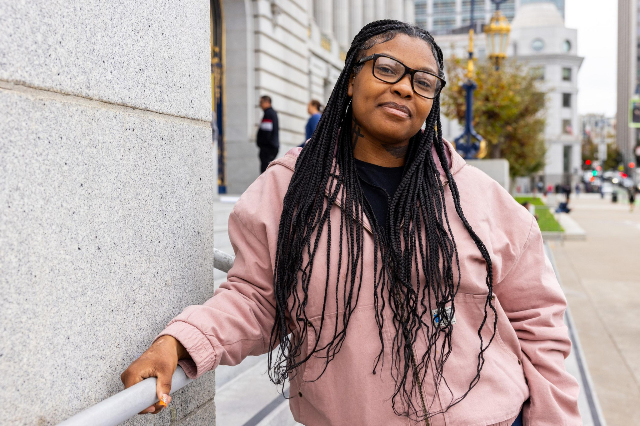 A woman with long braided hair and glasses leans against a railing outside a stone building, wearing a light pink jacket and a relaxed expression.
