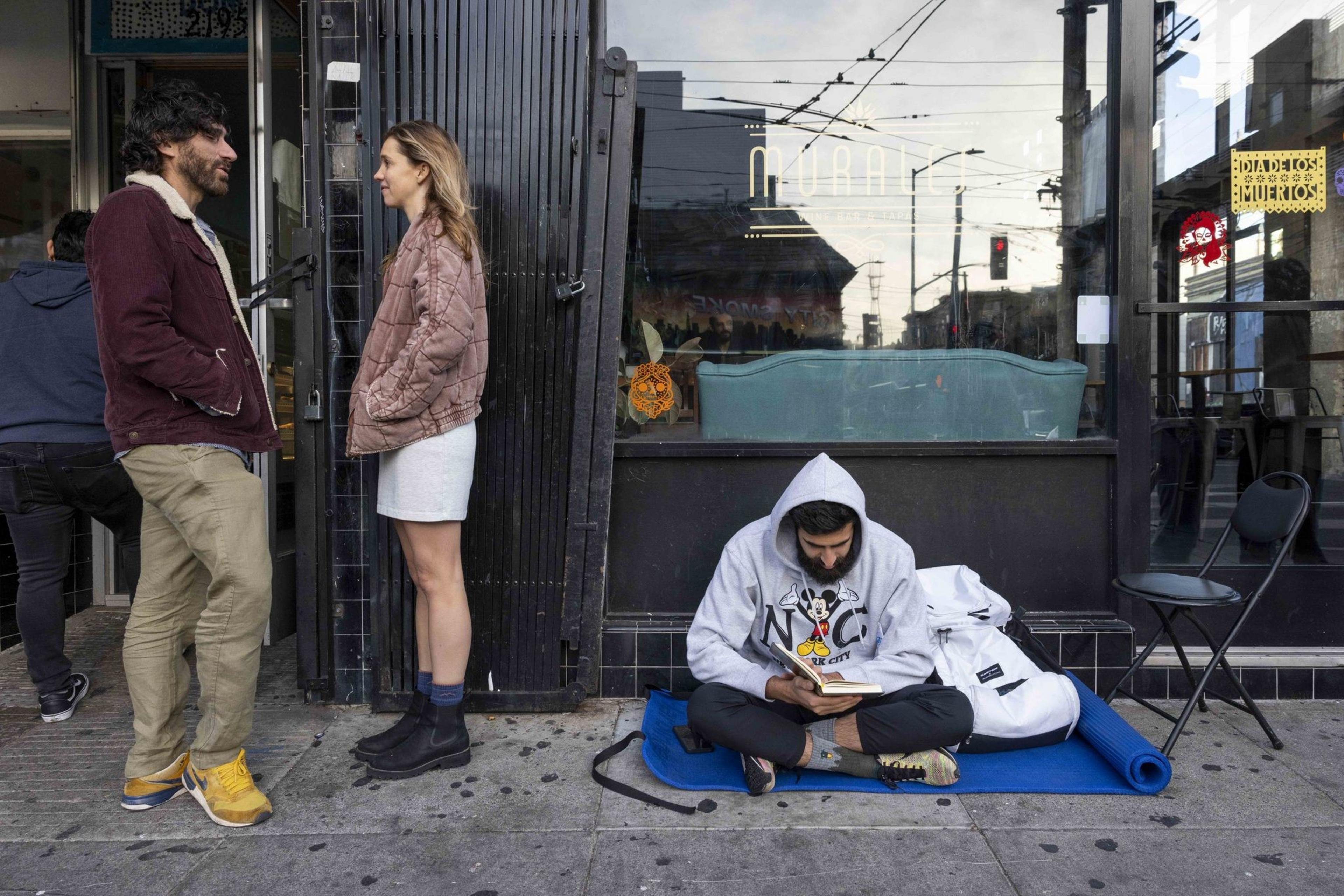 A man and woman stand outside a shop talking, while another man sits on the ground reading a book. He's wearing a hoodie and is surrounded by a mat and bag.
