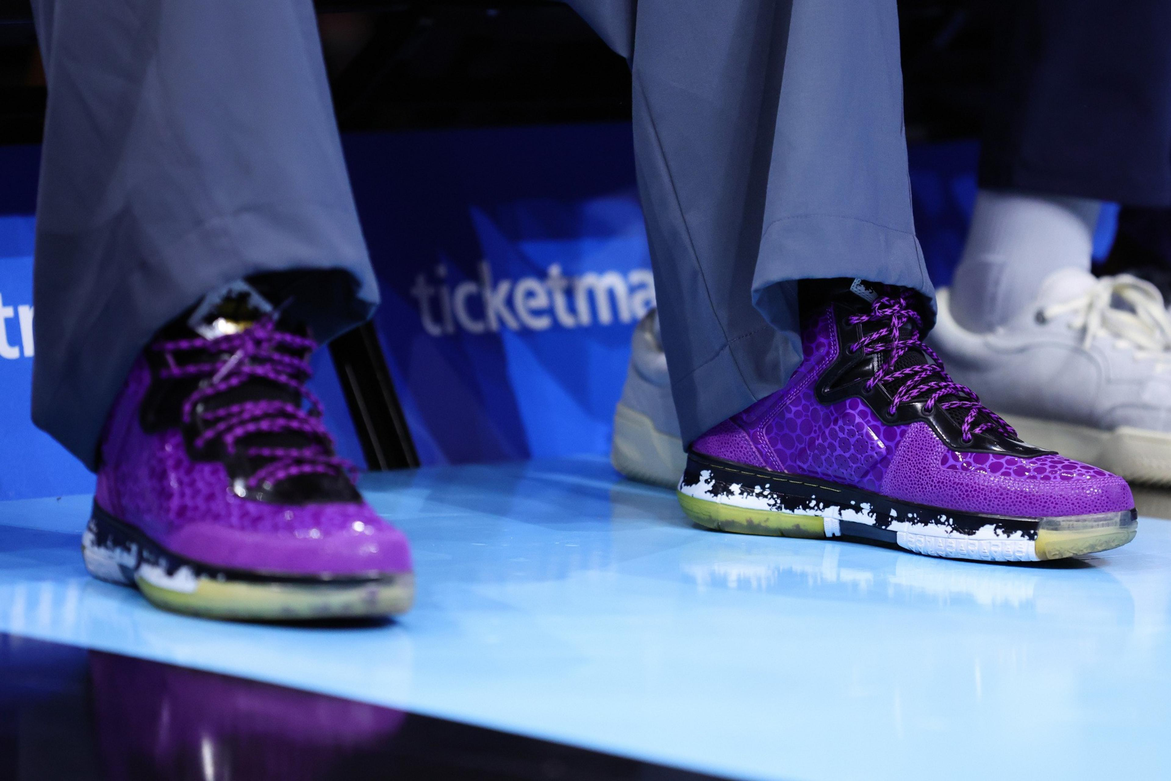 Purple high-top sneakers with black and white soles are worn with gray pants, sitting on a shiny blue floor next to someone in white shoes.