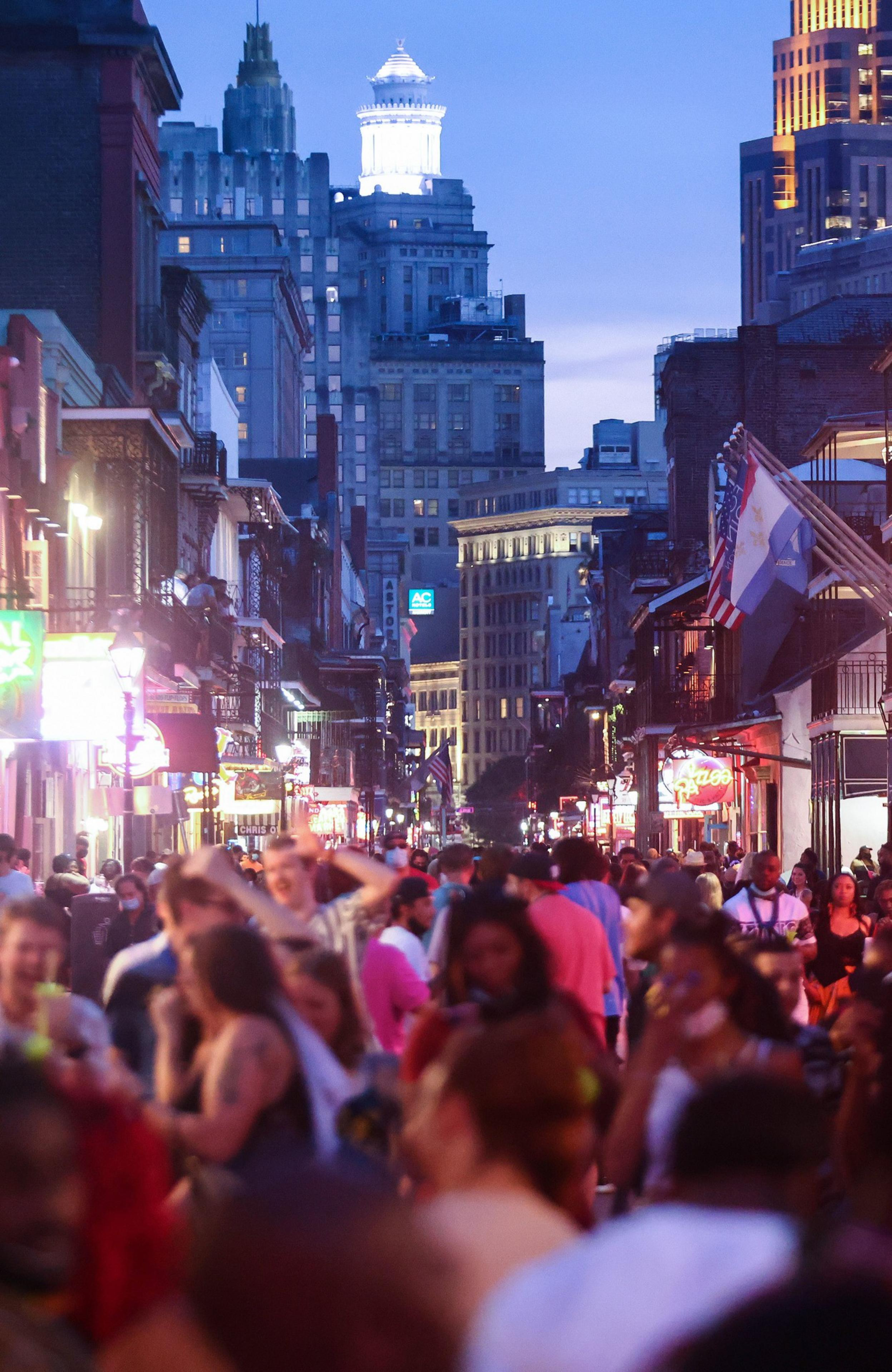 A bustling city street at dusk, filled with a diverse crowd and illuminated by neon signs, with a lit tower in the background.