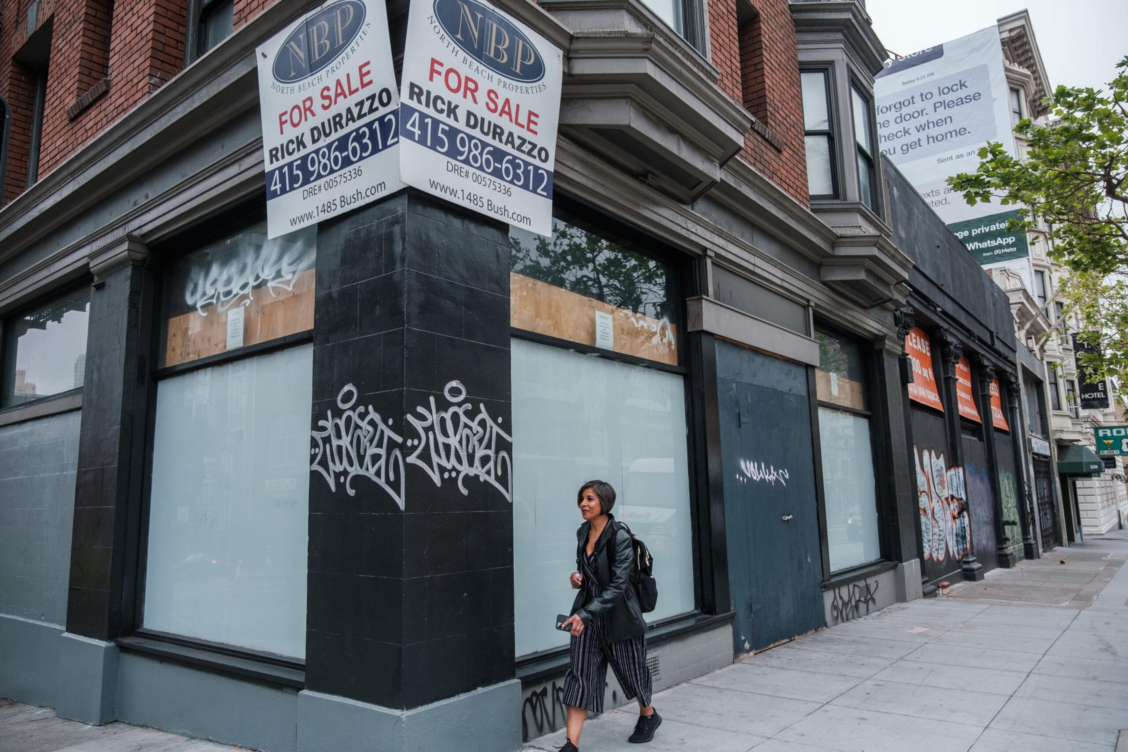 A woman walks past a corner building with boarded-up windows, graffiti, and “For Sale” signs advertising the property.