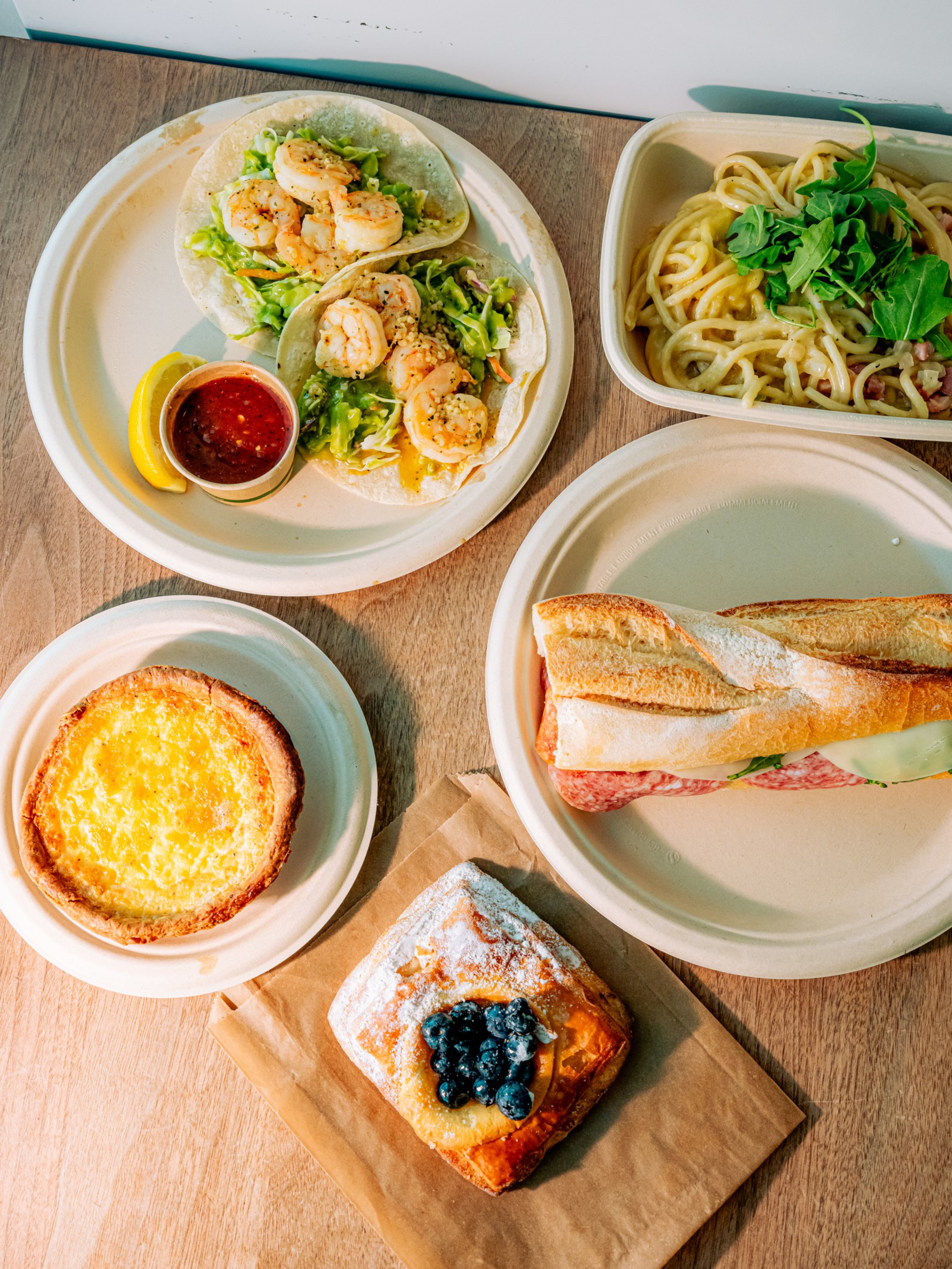 Two shrimp tacos with lettuce and salsa, a container of pasta with greens, a sandwich baguette with meat and cheese, a small quiche, and a pastry with blueberries.