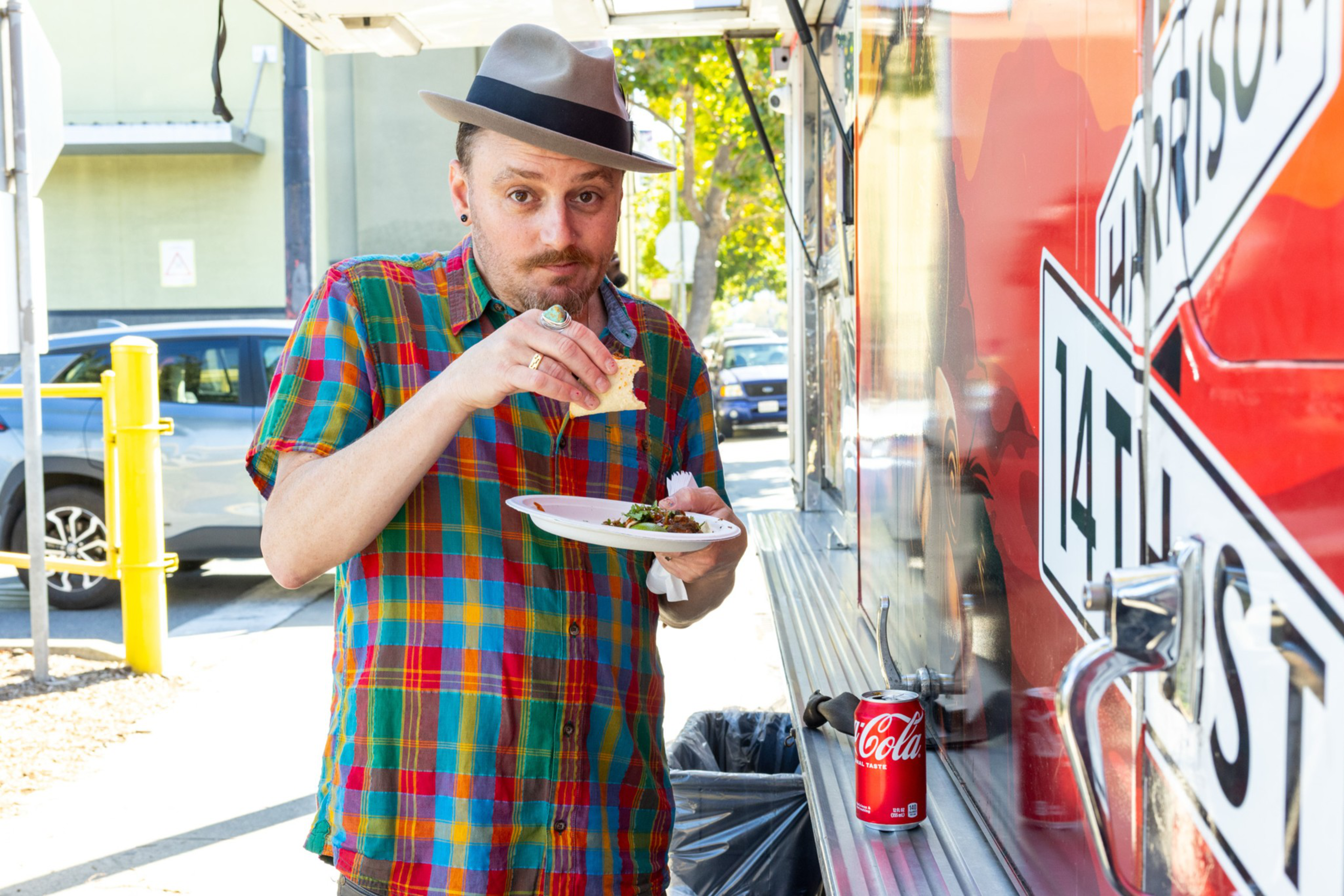 A man in a colorful plaid shirt and gray hat holds a plate and eats a taco while standing beside a red food truck with a can of Coke nearby.