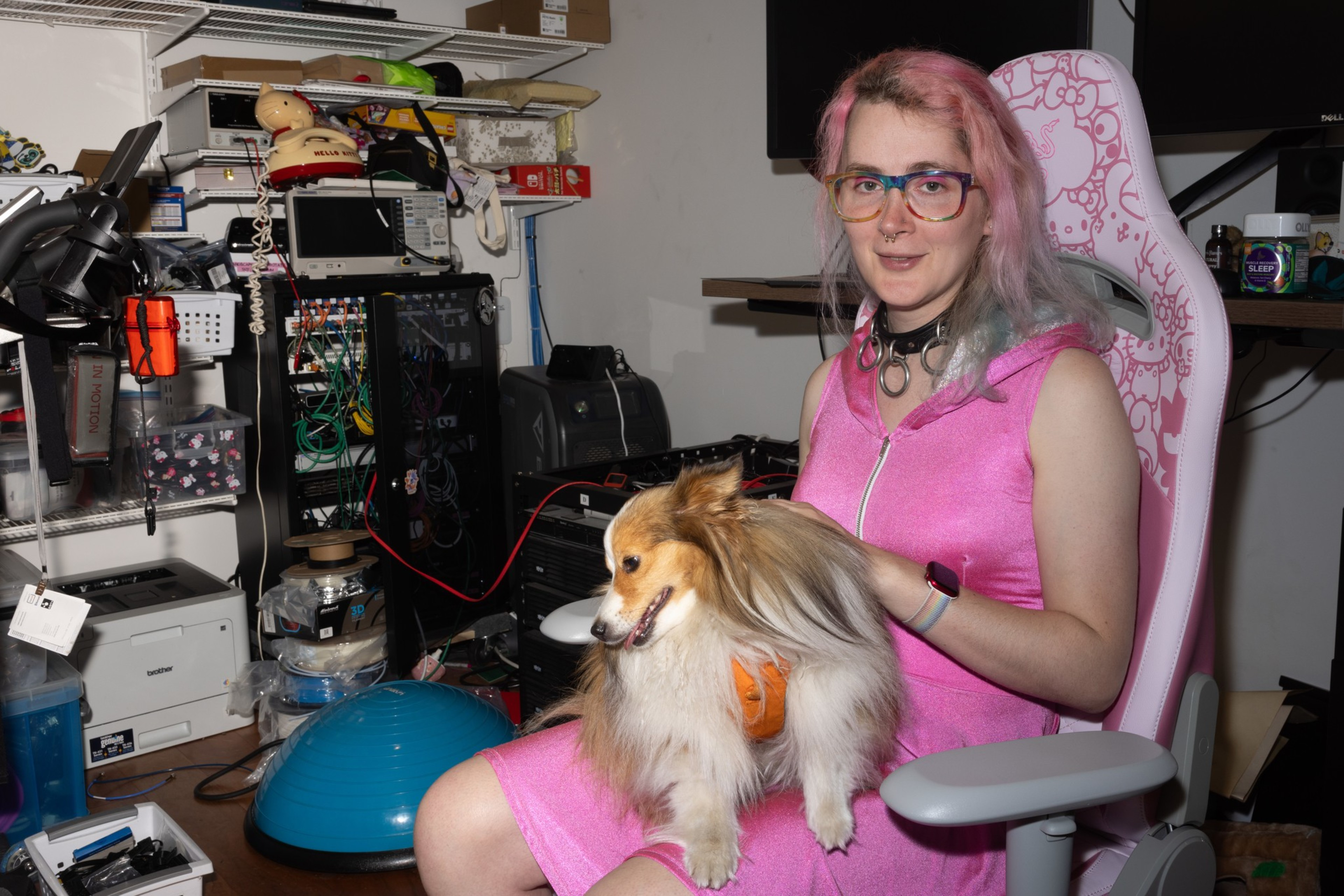 A person with pink hair sits in a white chair, wearing a pink dress and glasses, holding a small dog. The room is cluttered with electronics and various equipment.