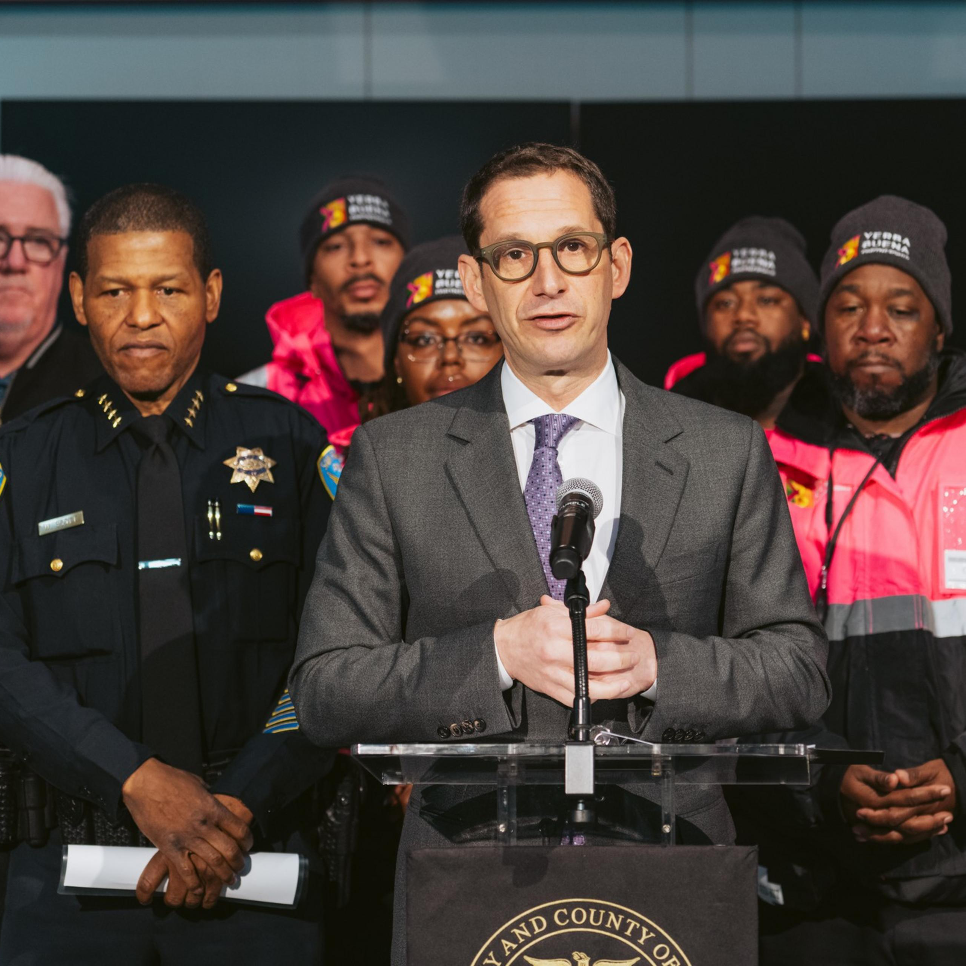A man in a suit speaks at a podium, surrounded by uniformed officers and individuals in bright pink jackets, likely indicating a formal announcement or briefing.