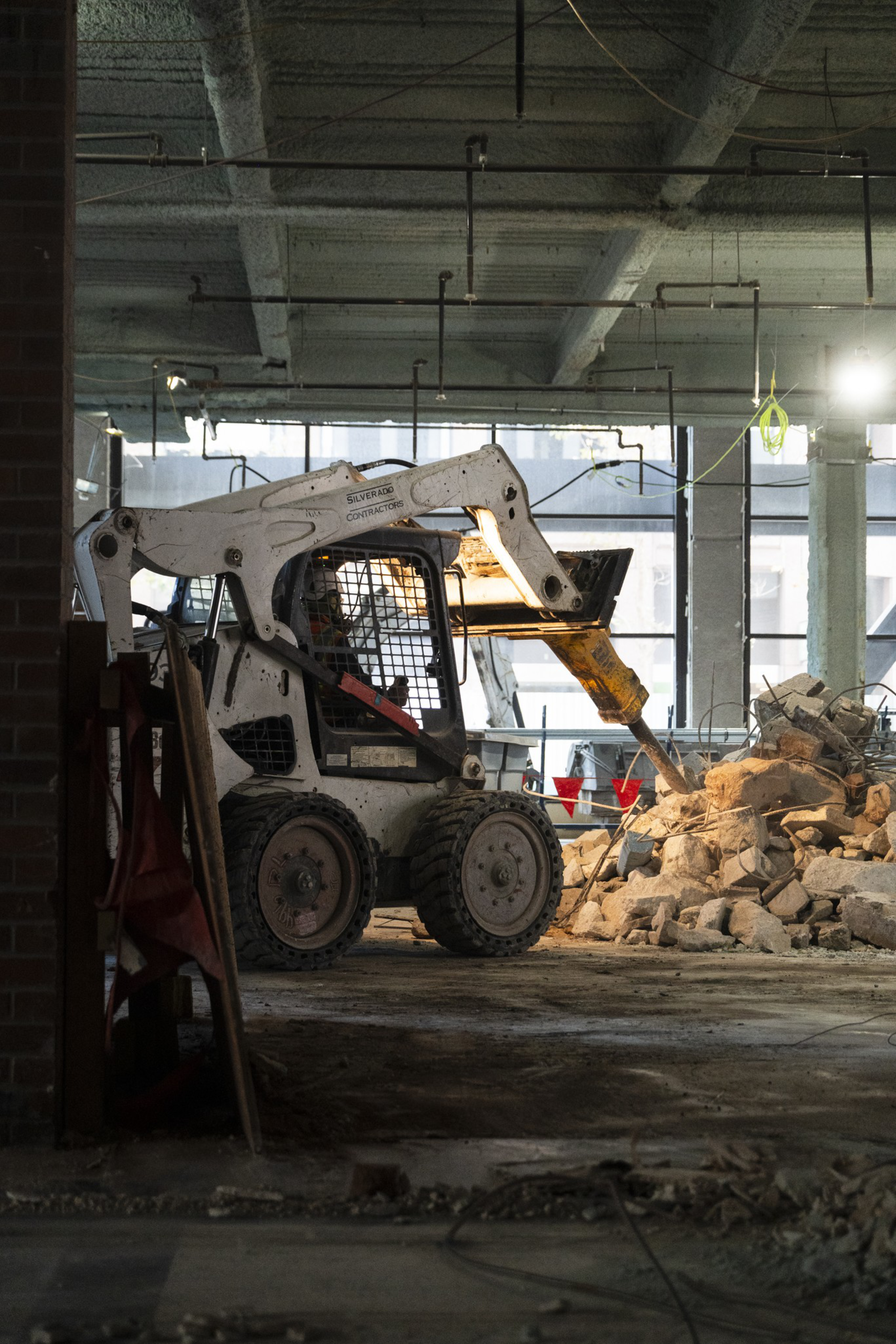 A small skid-steer loader with a hydraulic hammer attachment breaks concrete rubble inside a dimly lit, unfinished building.