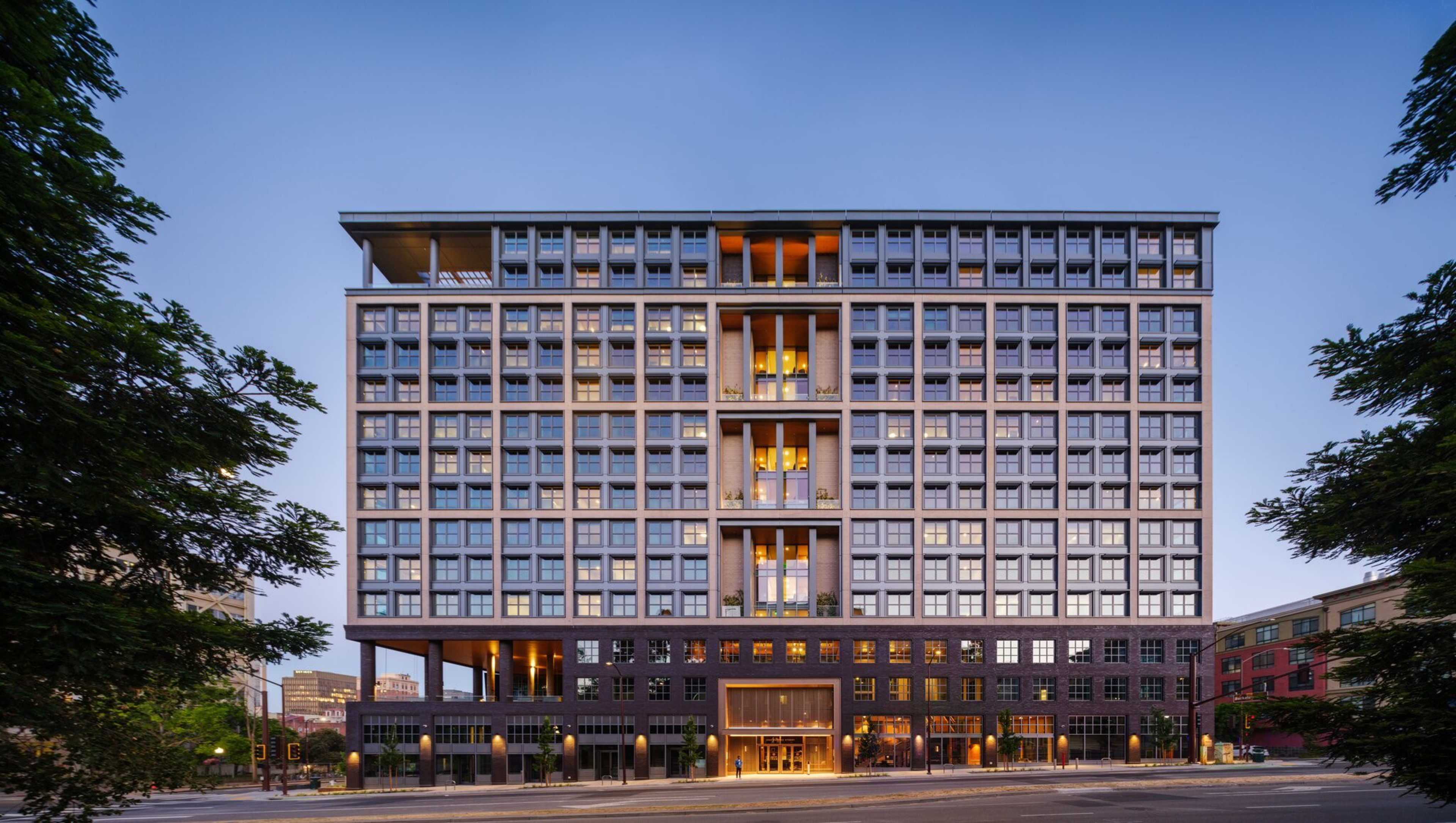 The image shows a modern, multi-story building with a grid-like window pattern and a mix of light and dark facade elements, set against a clear sky. Trees frame the sides.