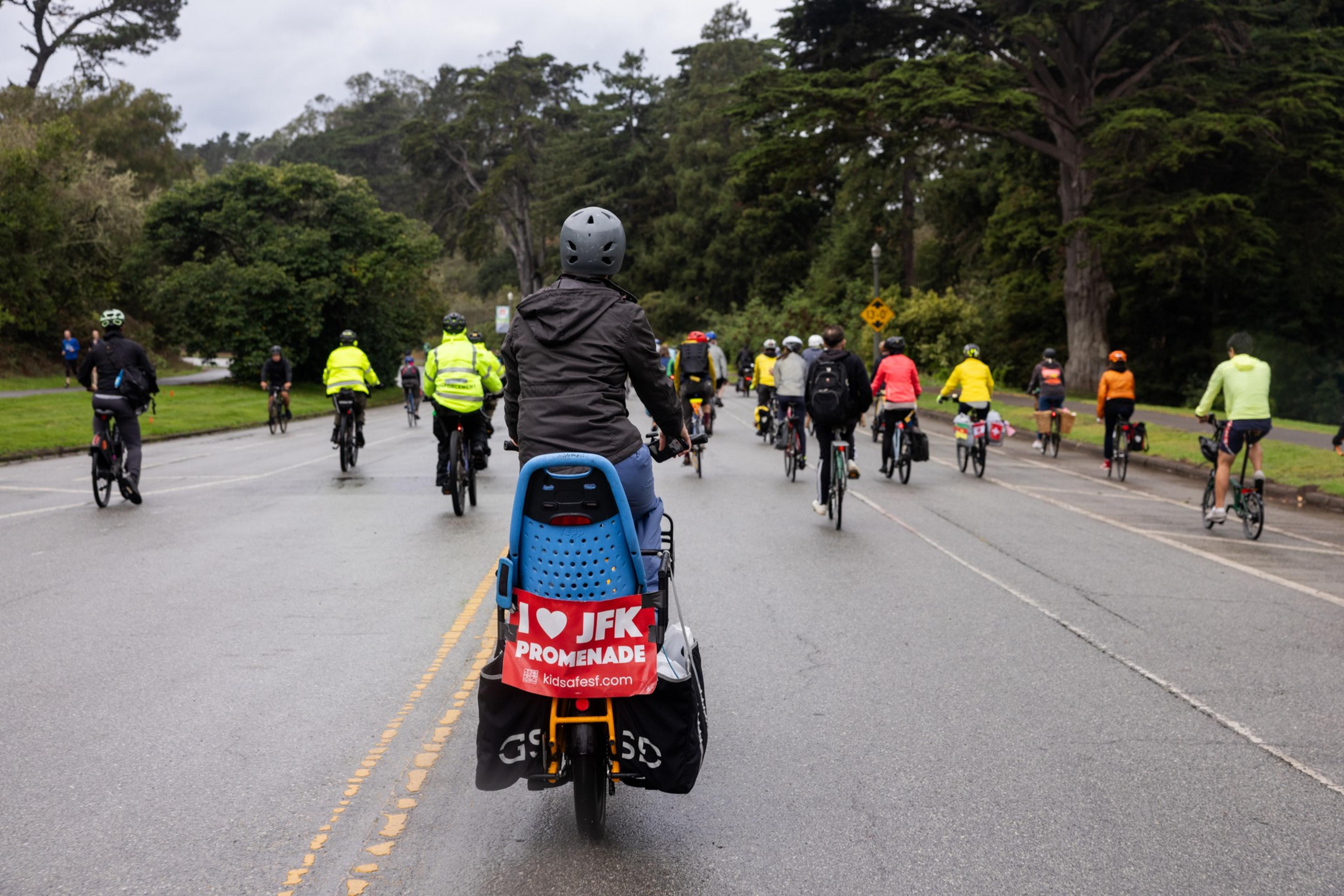 A group of people ride their bike through Golden Gate Park and on the back of one of the bikes hangs a sign that reads, &quot;I Love JFK Promenade.