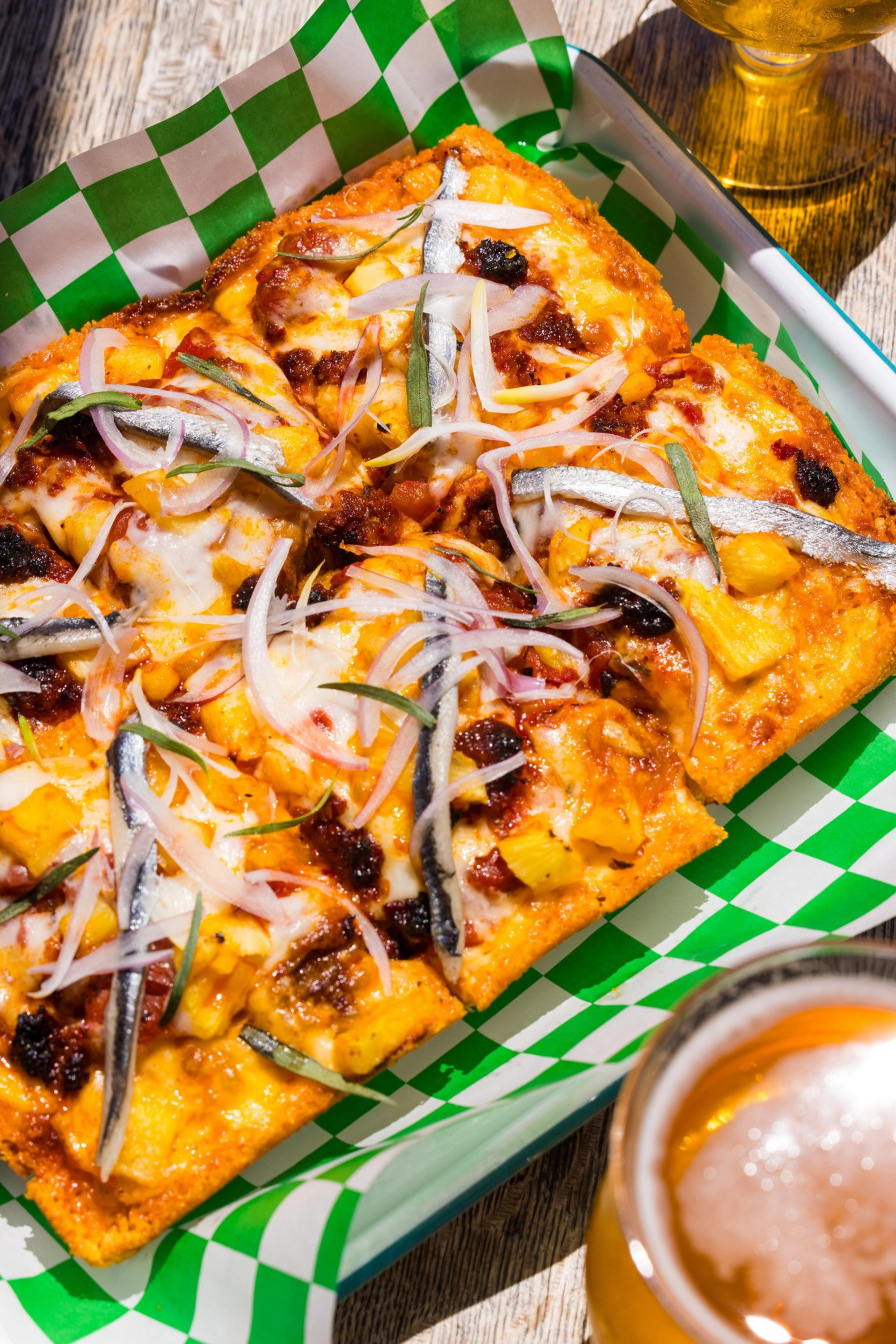 A rectangular pizza topped with anchovies, onions, cheese, and sauce rests on a green-checkered paper next to a glass of frothy beer.