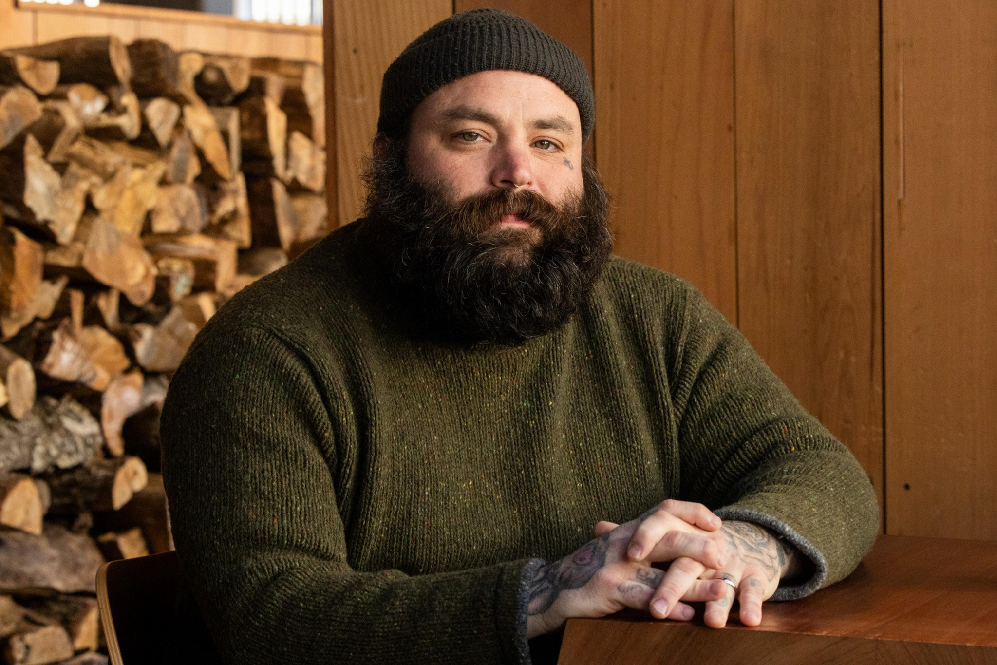 A bearded man in a beanie and sweater sits at a wooden table, hands clasped. Behind him is a stack of cut logs against a wooden wall.