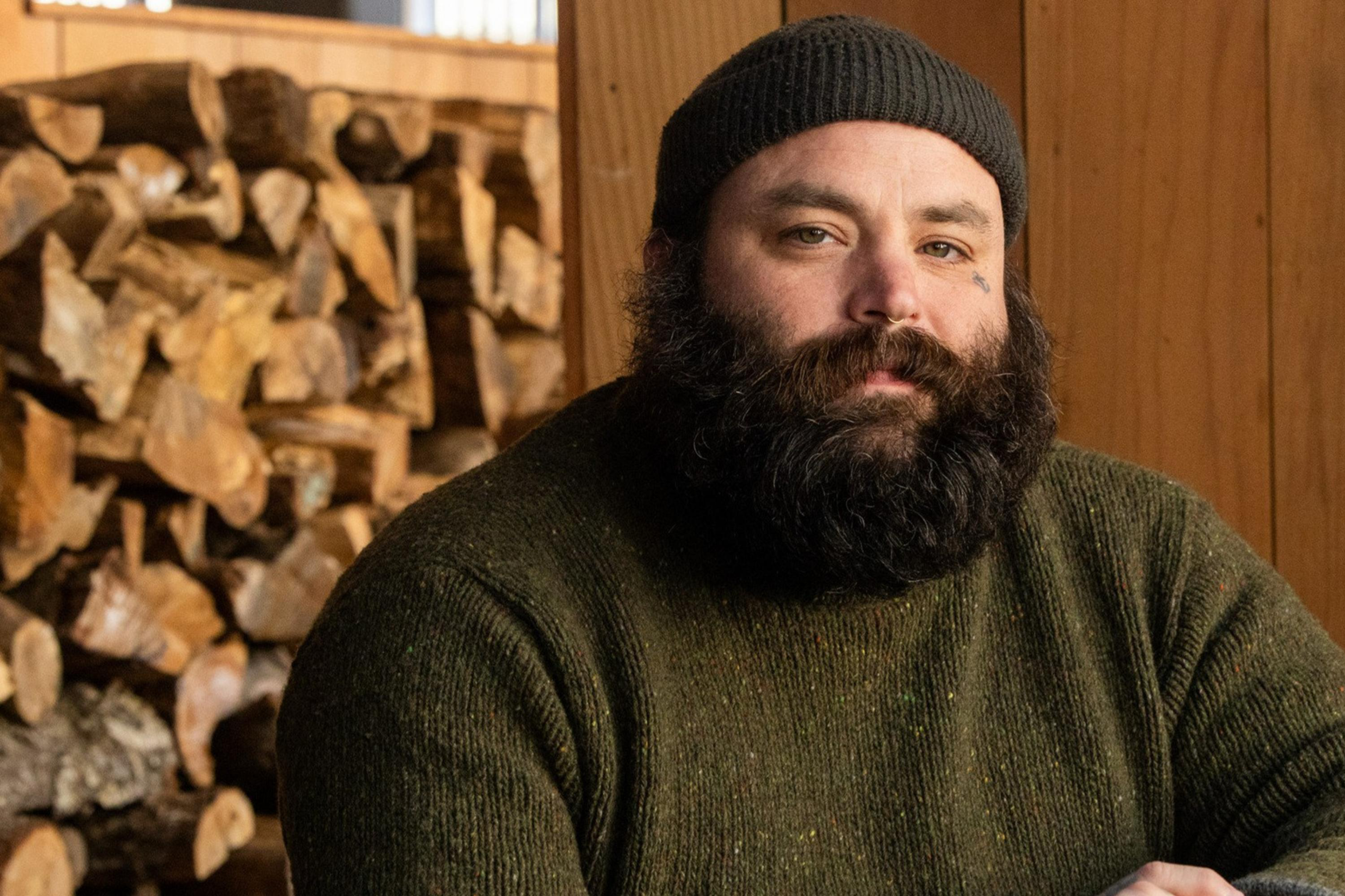 A bearded man in a beanie and sweater sits at a wooden table, hands clasped. Behind him is a stack of cut logs against a wooden wall.