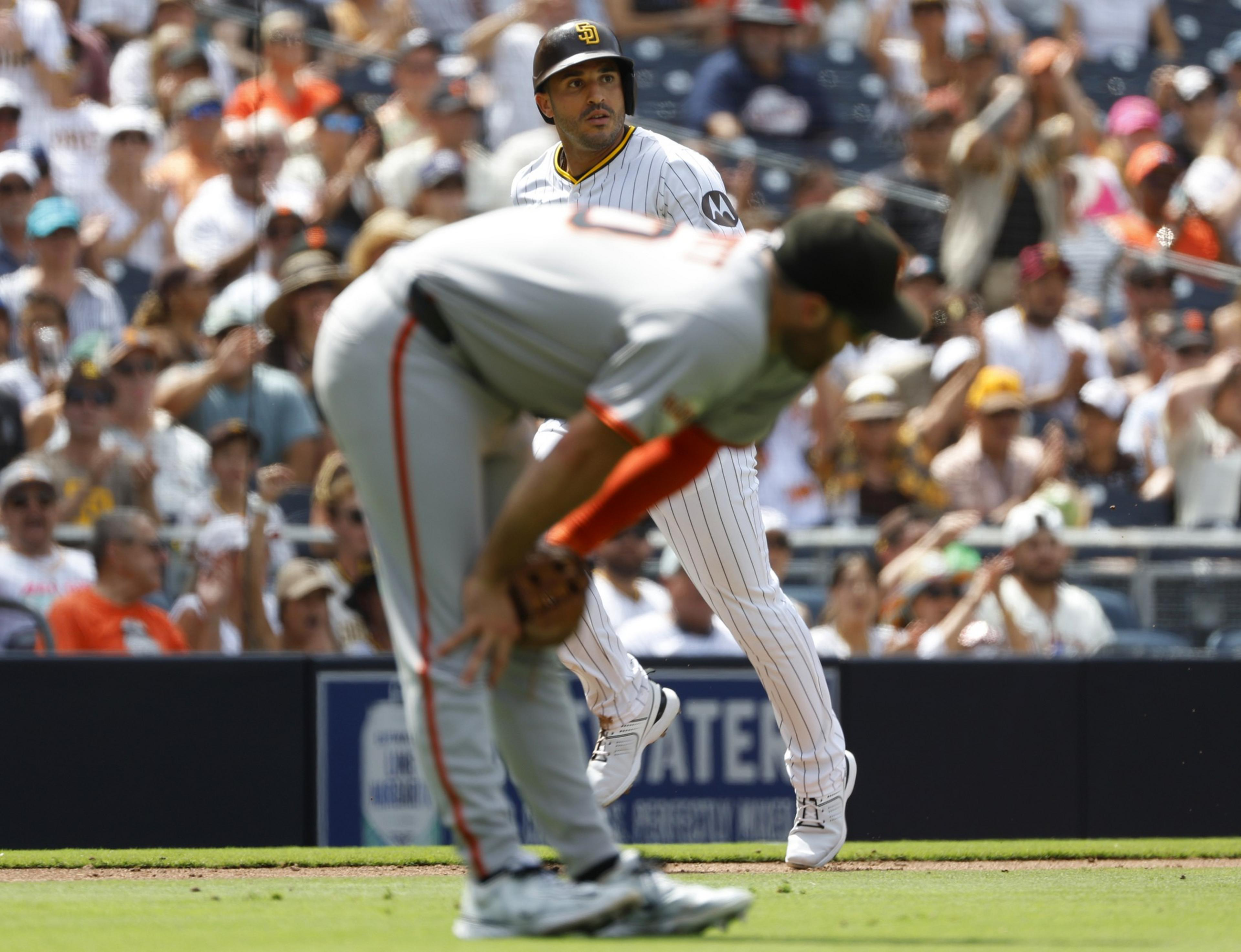 A baseball player in a white pinstripe uniform looks intently while an opposing player in gray bends over in the foreground with a crowd watching behind.
