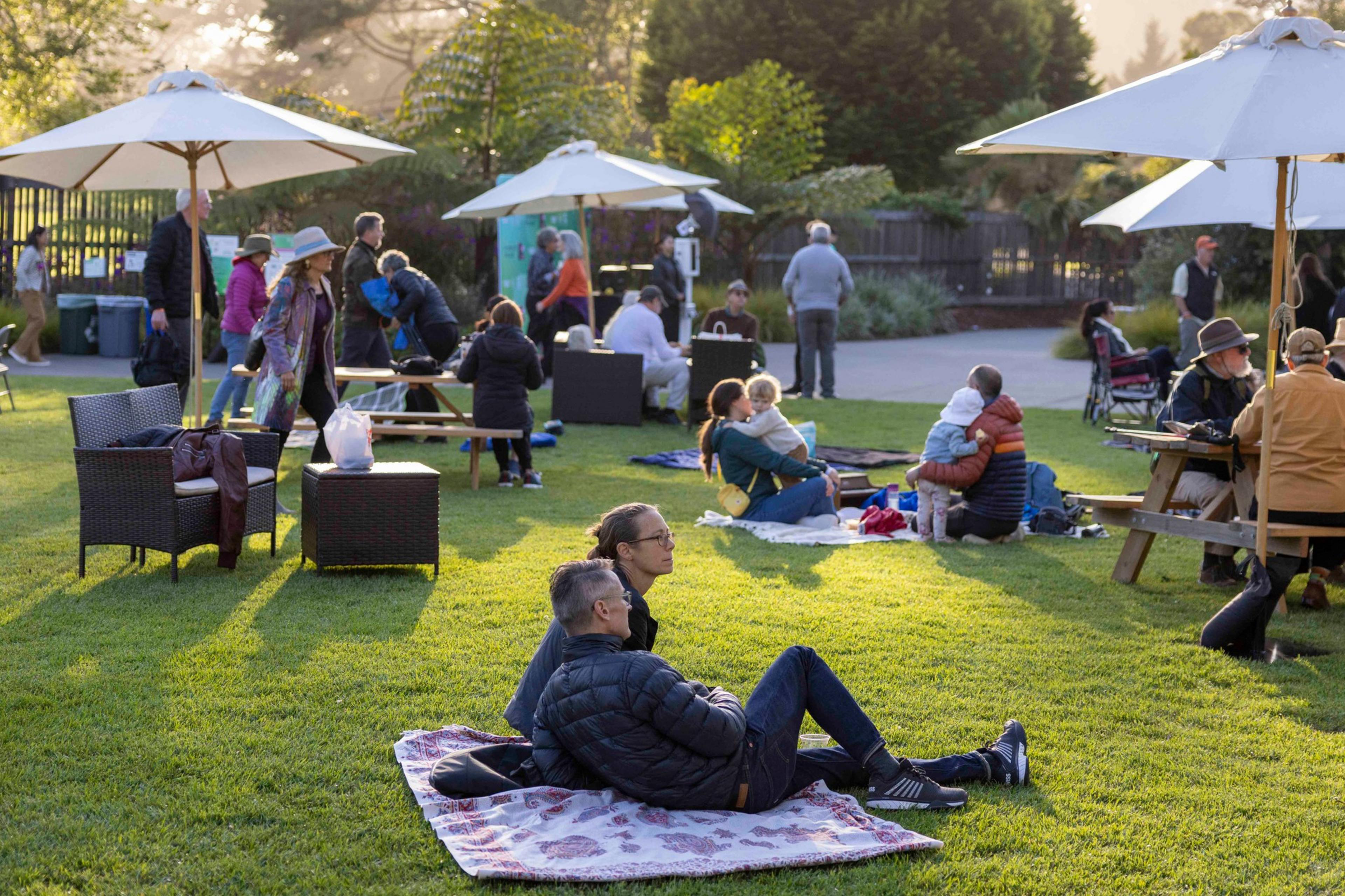 People are relaxing on a grassy area with blankets, wicker chairs, and picnic tables under sun umbrellas, enjoying a park-like setting in the late afternoon.