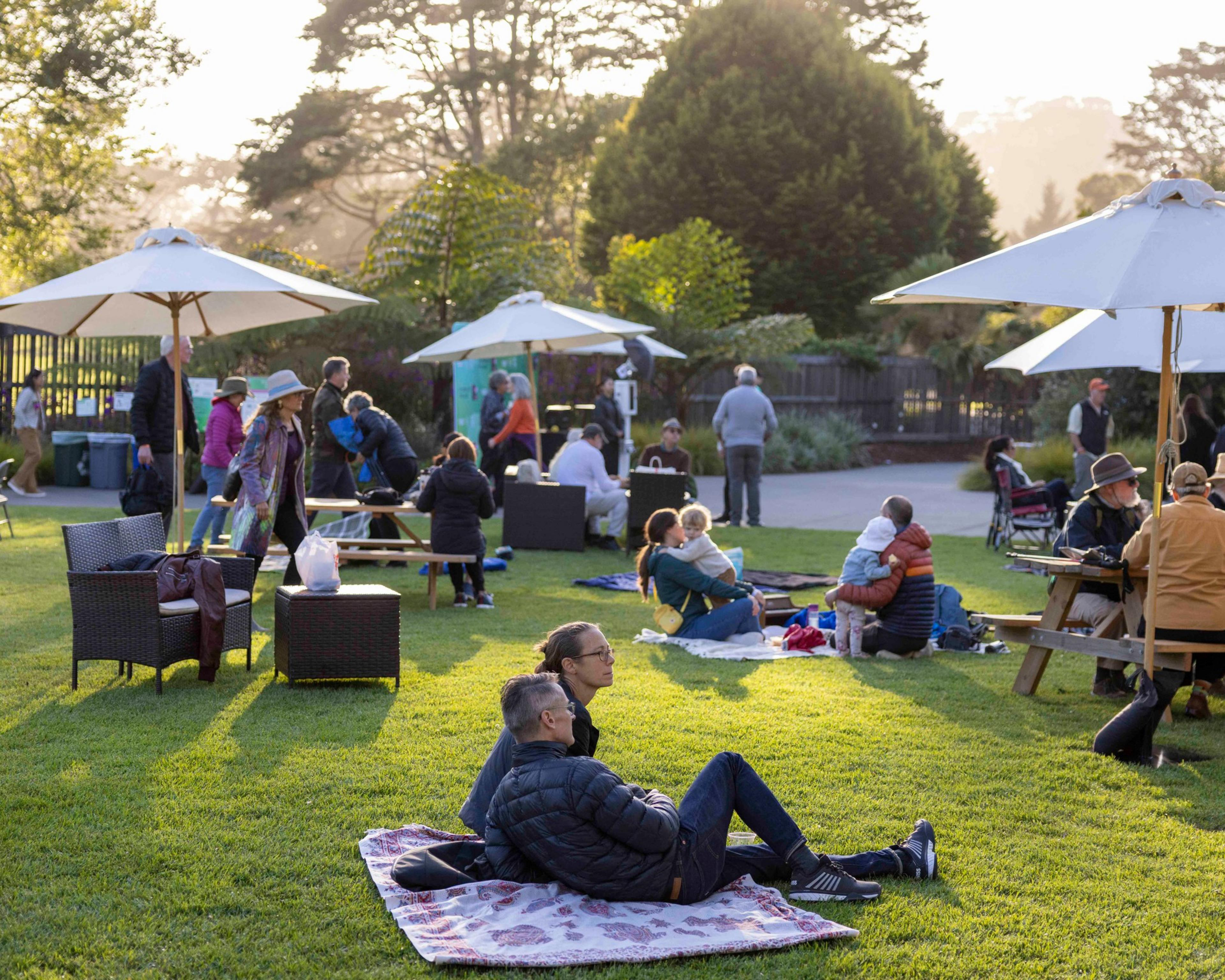 People are relaxing on a grassy area with blankets, wicker chairs, and picnic tables under sun umbrellas, enjoying a park-like setting in the late afternoon.