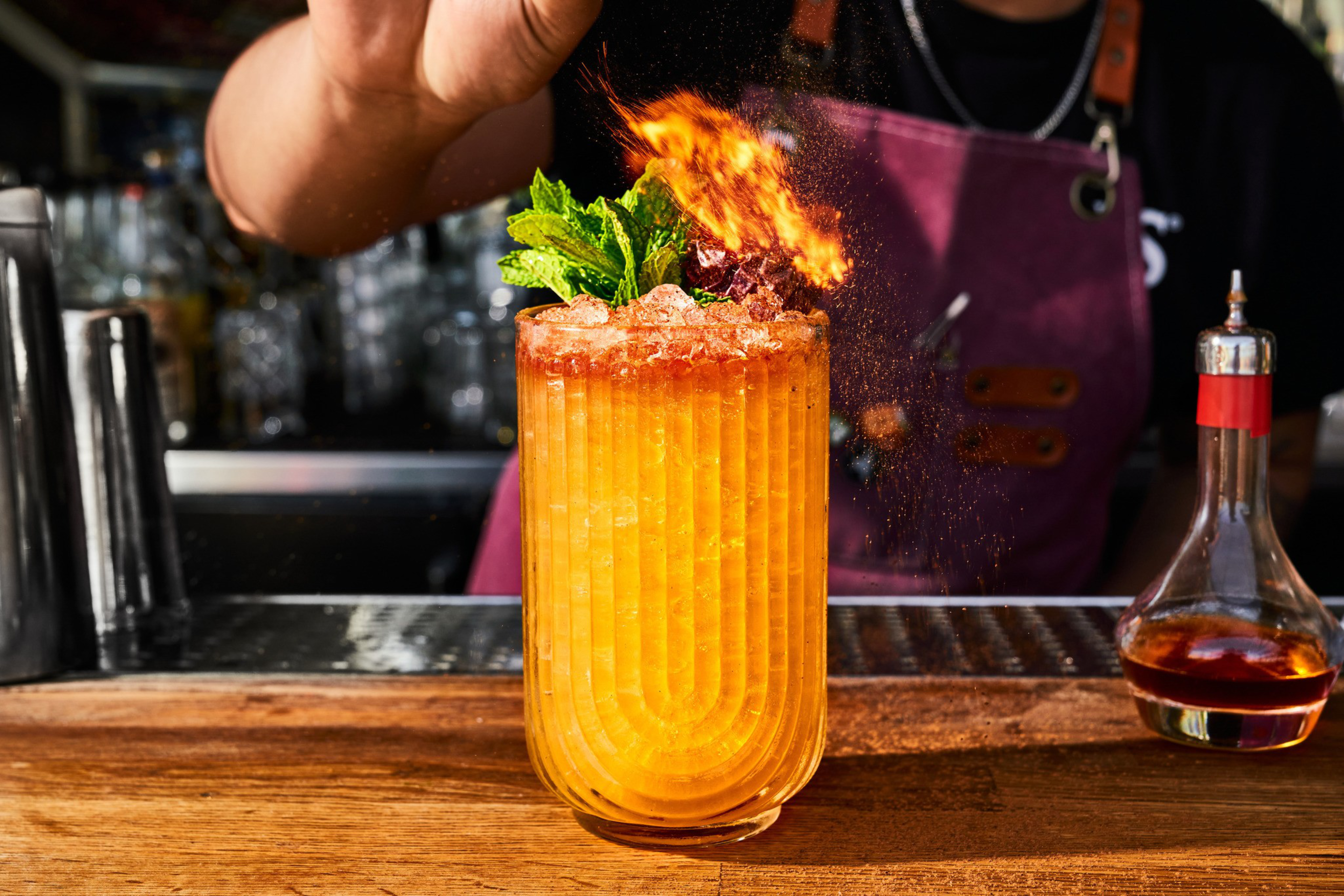 A vibrant orange cocktail in a ribbed glass is garnished with mint and flaming spices. It's set on a bar, with a person in a pink apron behind it.