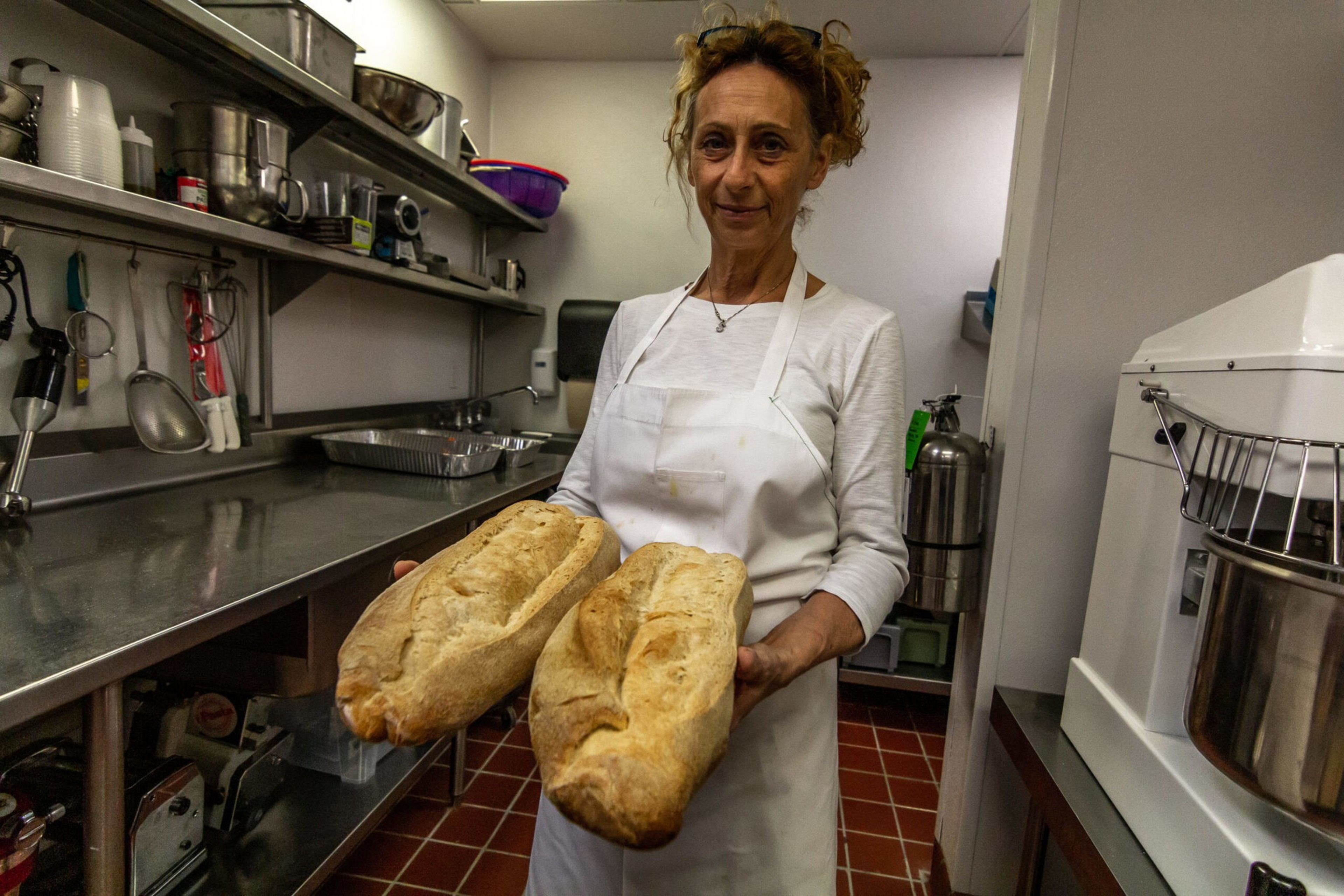 A woman wearing a white apron holds two large loaves of freshly baked bread in a commercial kitchen.