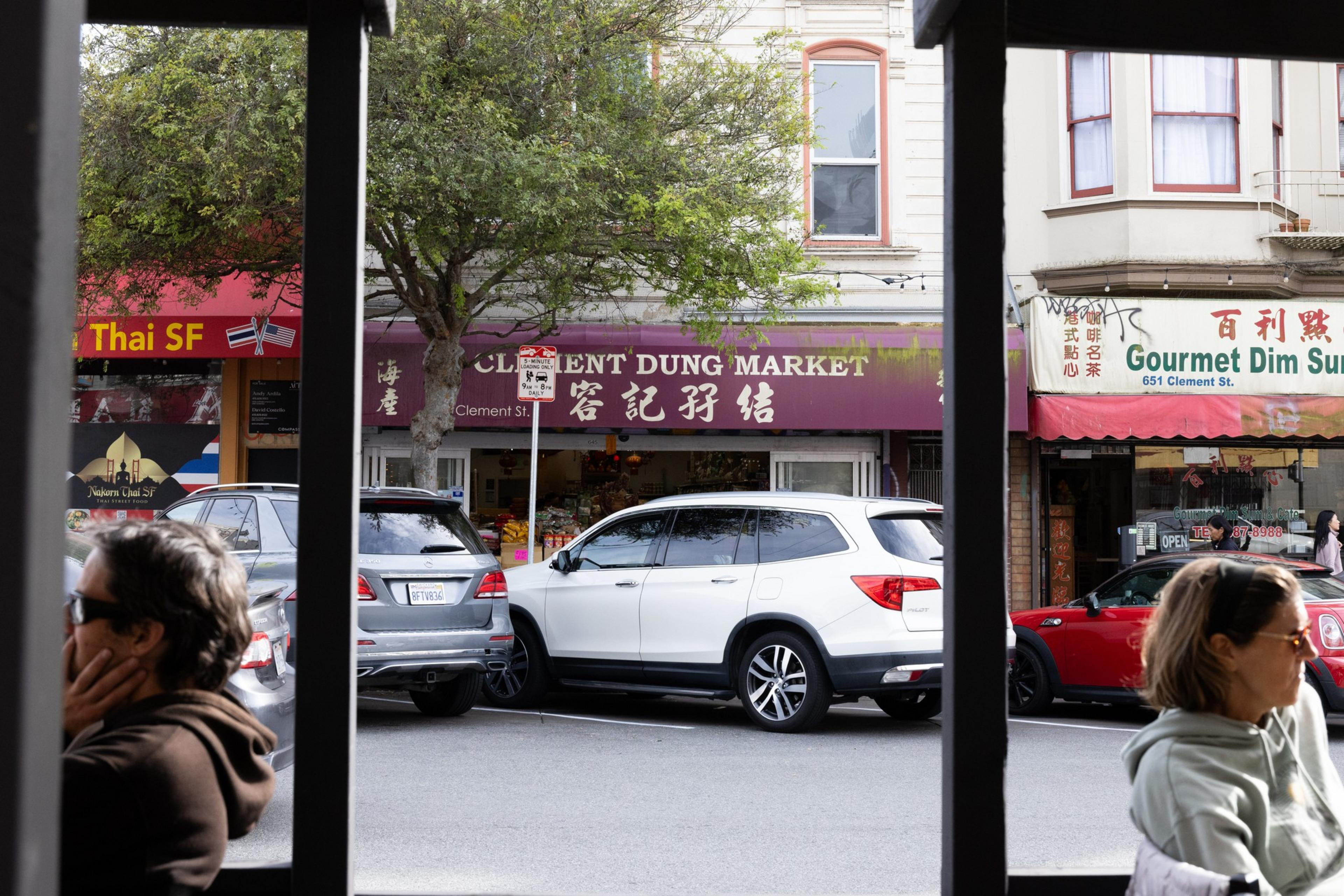 People sitting outside, viewing a street with parked cars in front of Asian markets and restaurants, like "Clement Dung Market" and "Gourmet Dim Sum."