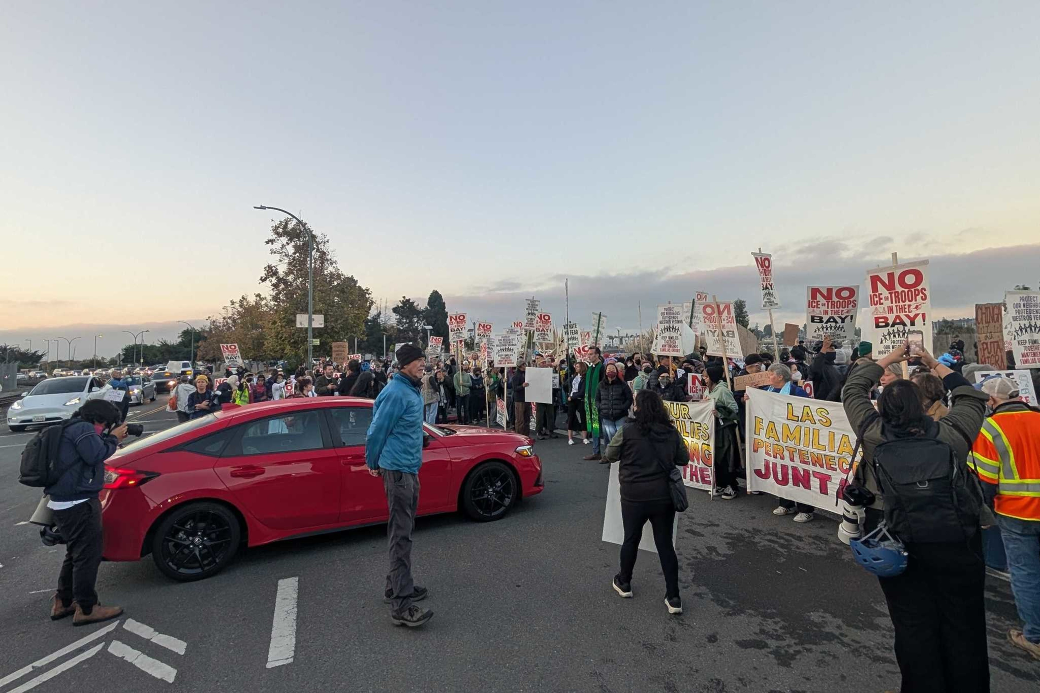 A large crowd of protesters holds signs opposing ICE troops and advocating for family unity near a red car blocking part of the street.