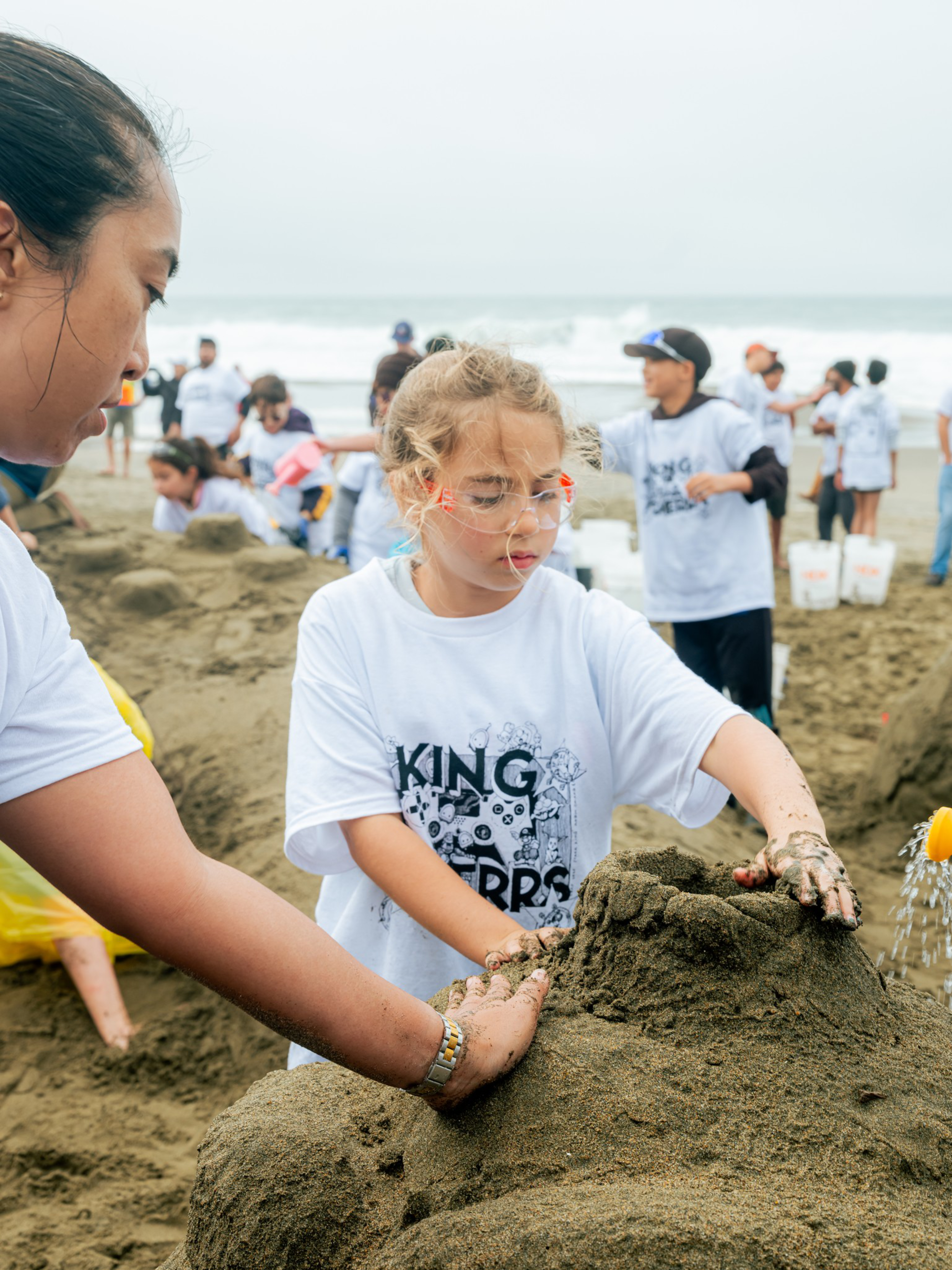 Children wearing white shirts with “King” on them build sandcastles on a beach with the ocean and more participants in the background.