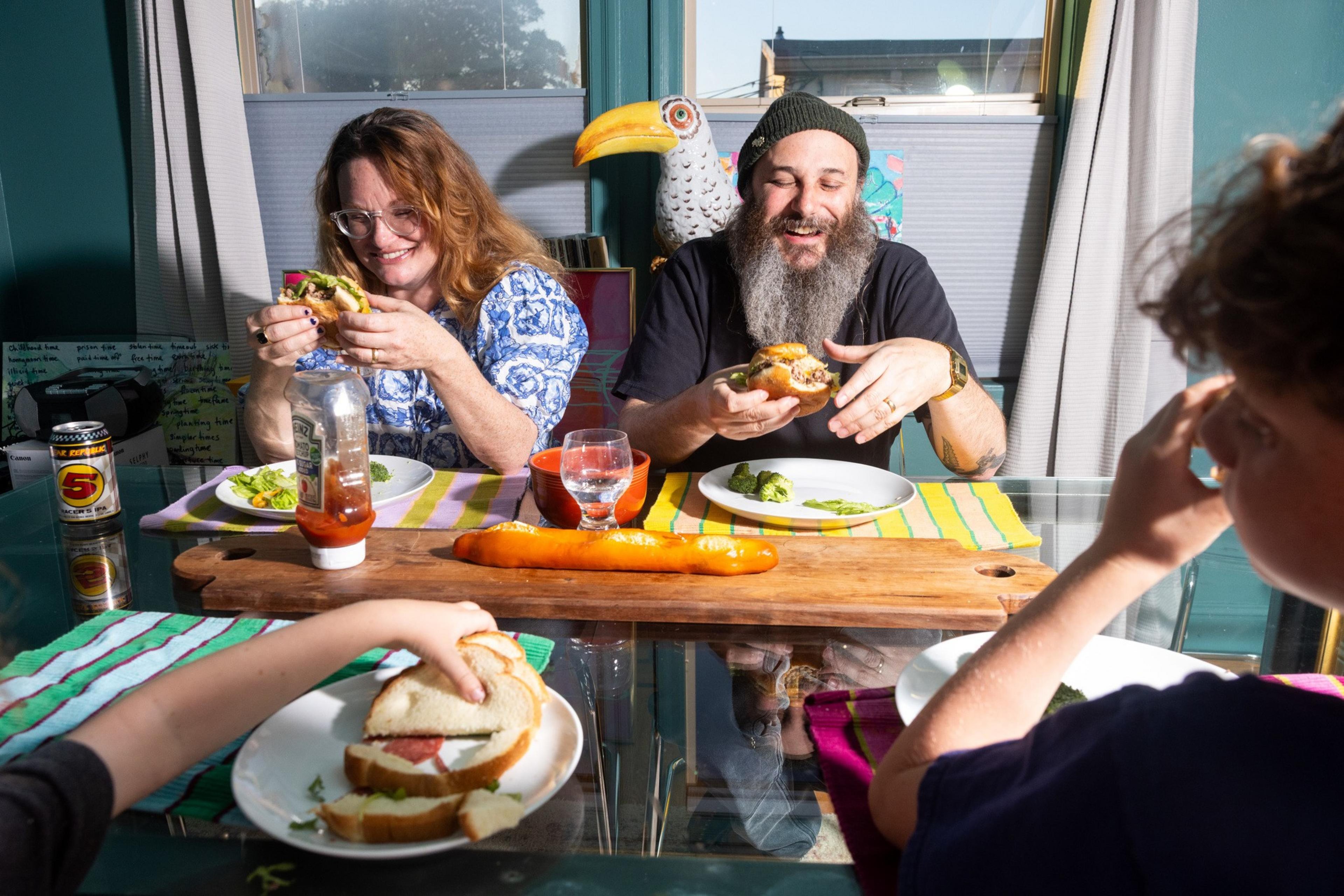 A family of three is joyfully eating sandwiches at a table. The table has a wooden platter, a bottle of ketchup, broccoli, and a can of soda.