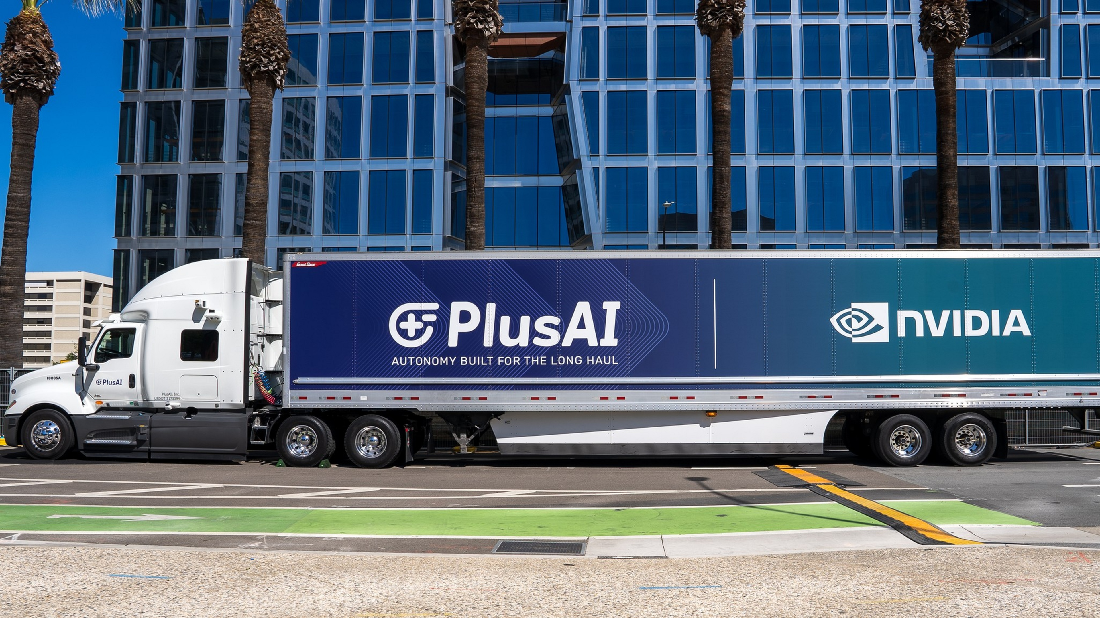 A white semi truck with a trailer displaying "PlusAI" and "NVIDIA" logos and the slogan "Autonomy built for the long haul" parked in front of a glass building.