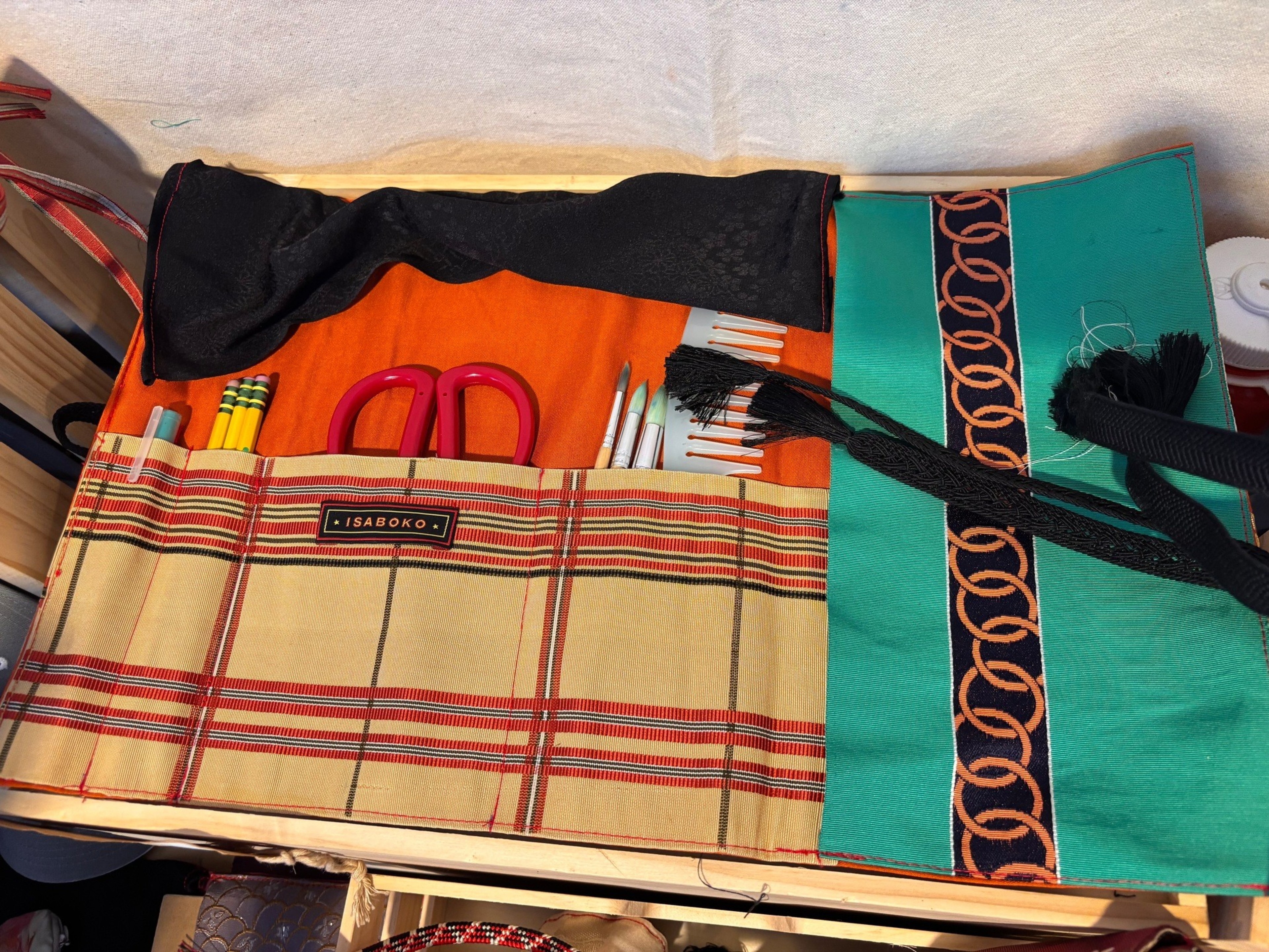 A fabric roll-up case with multiple pockets holds pencils, paintbrushes, scissors, and combs, featuring a plaid pattern and the label “ISABOKO.”