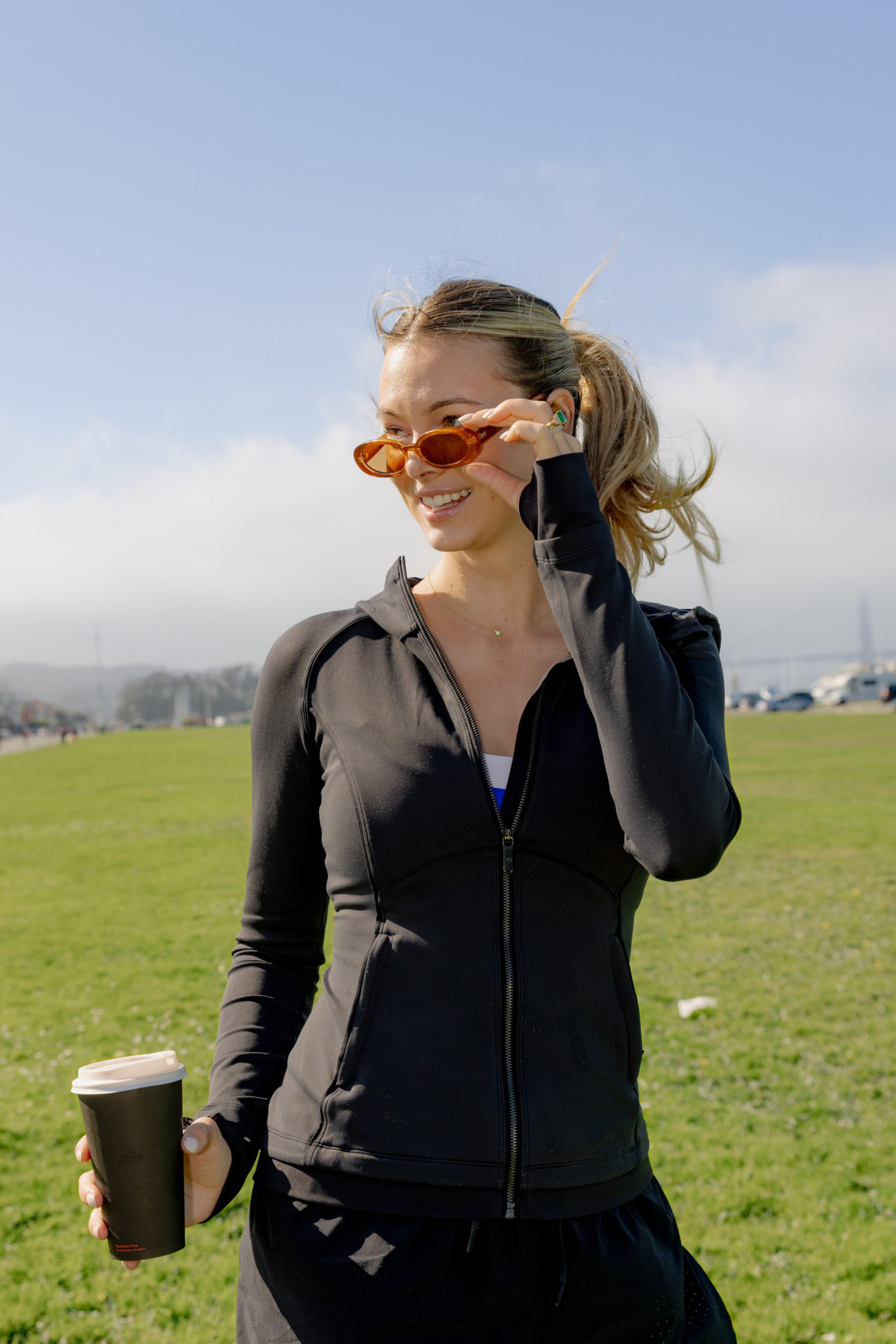 A woman in a black zip-up jacket and orange sunglasses smiles while holding a takeaway coffee cup outdoors on a sunny day with a grassy field behind her.