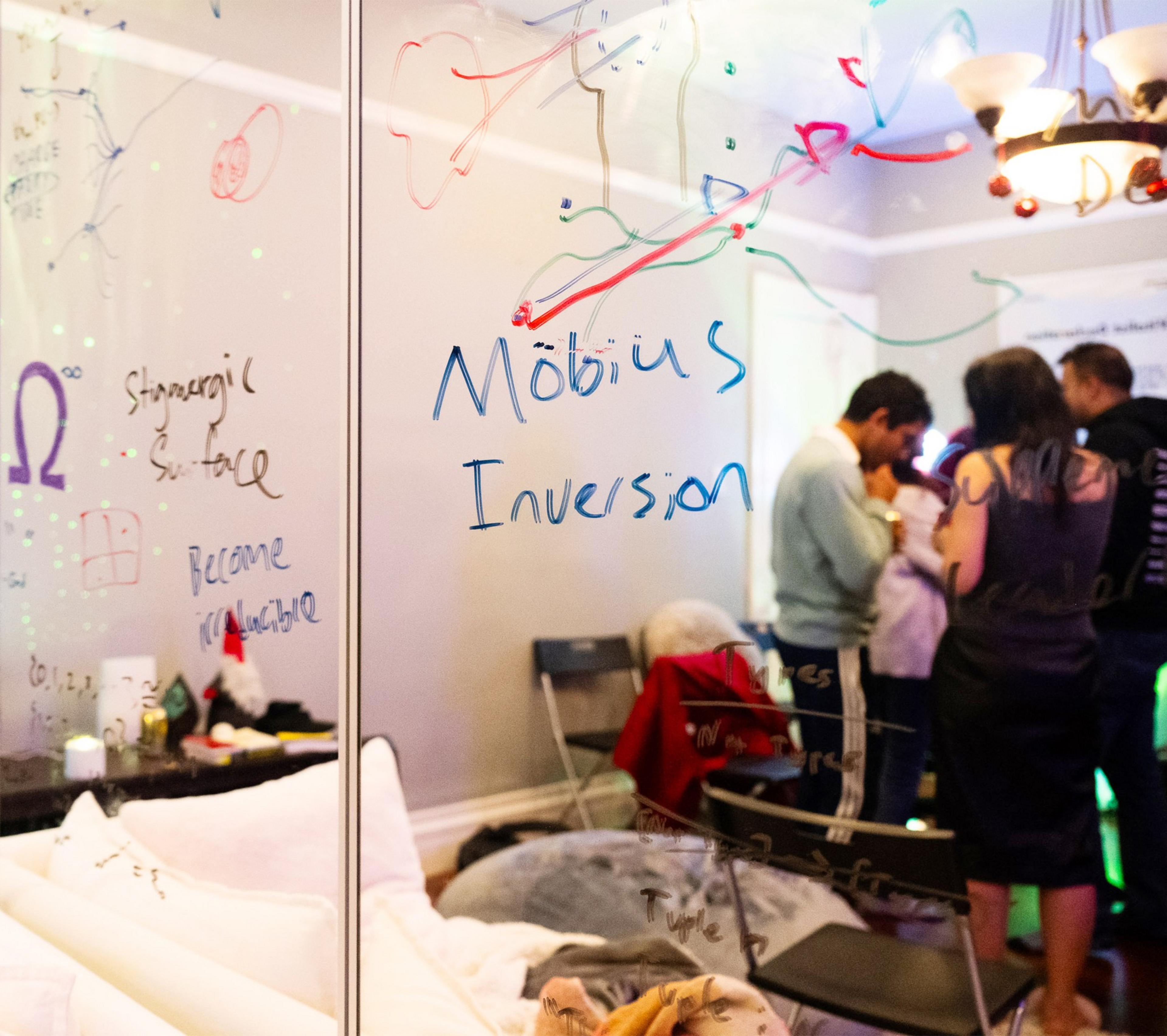 A room with people, a chandelier, and a glass board with colorful writings like &quot;Möbius Inversion.&quot; Chairs and items are scattered around.