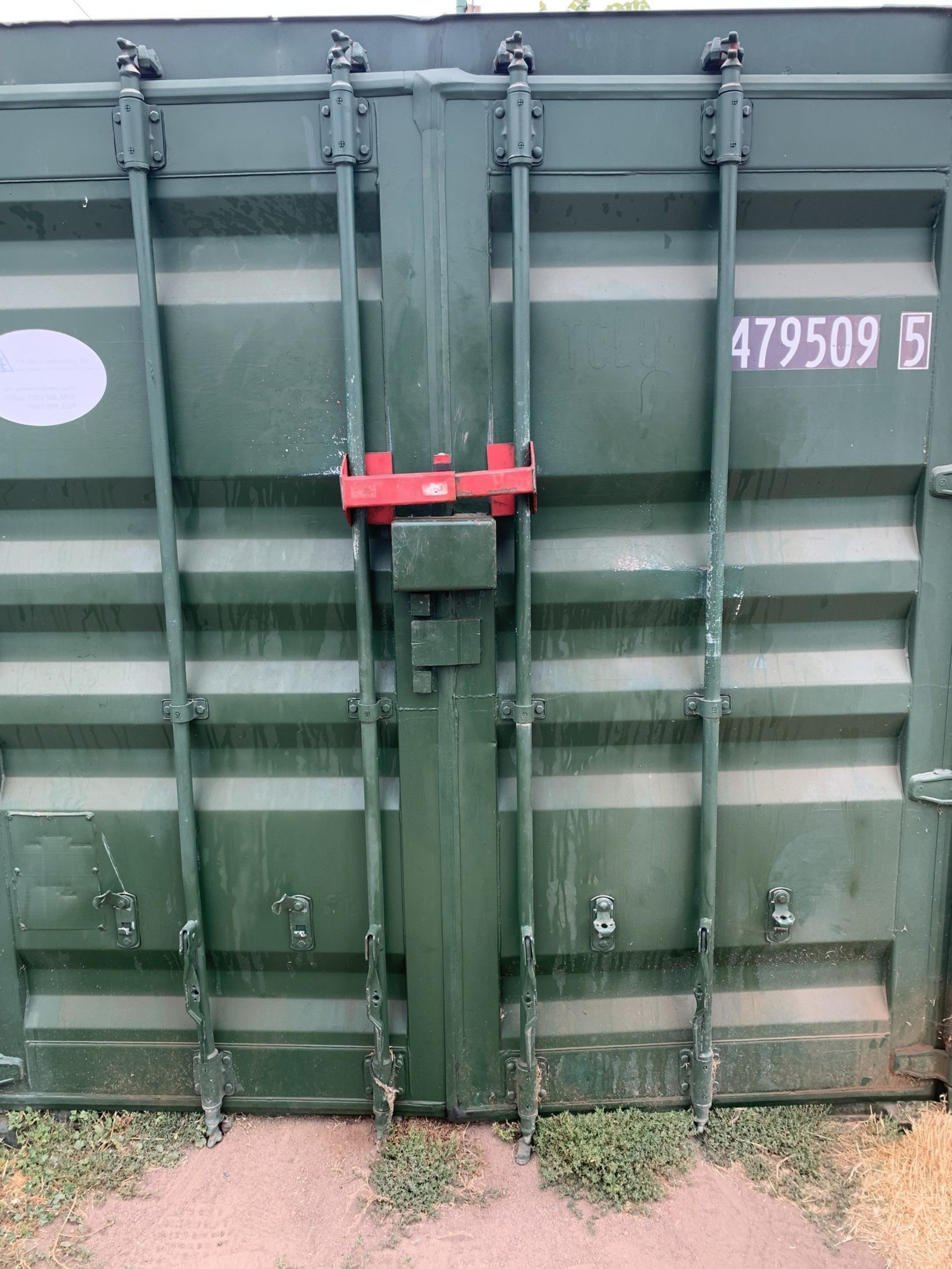 A green container door is secured with a thick red lock bar, featuring multiple latches and bolts. It has a number &quot;479509 5&quot; visible and sits on a grassy, sandy surface.