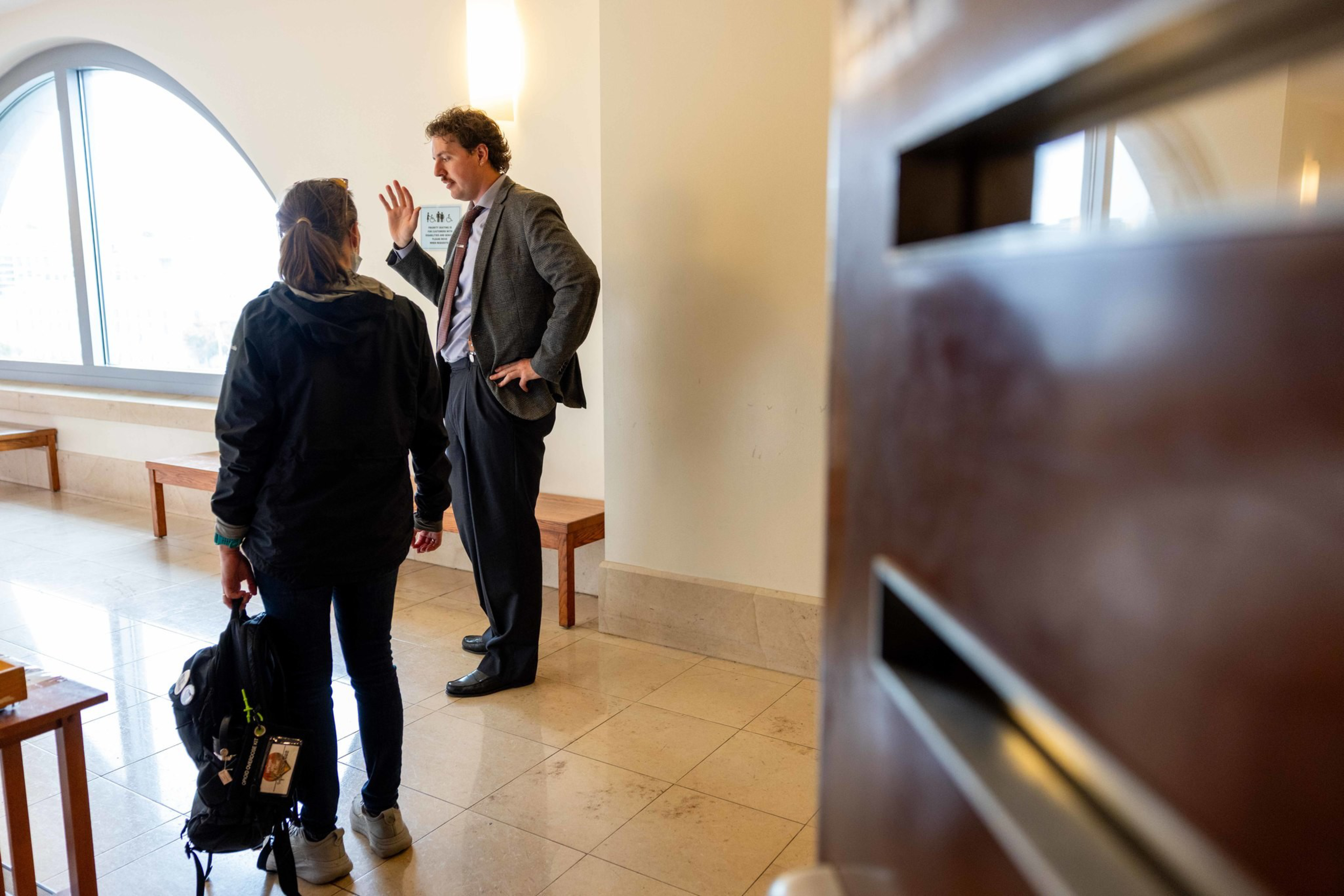 A man in a suit is talking to a woman wearing casual clothes and holding a backpack in a hallway with benches and a large arched window.