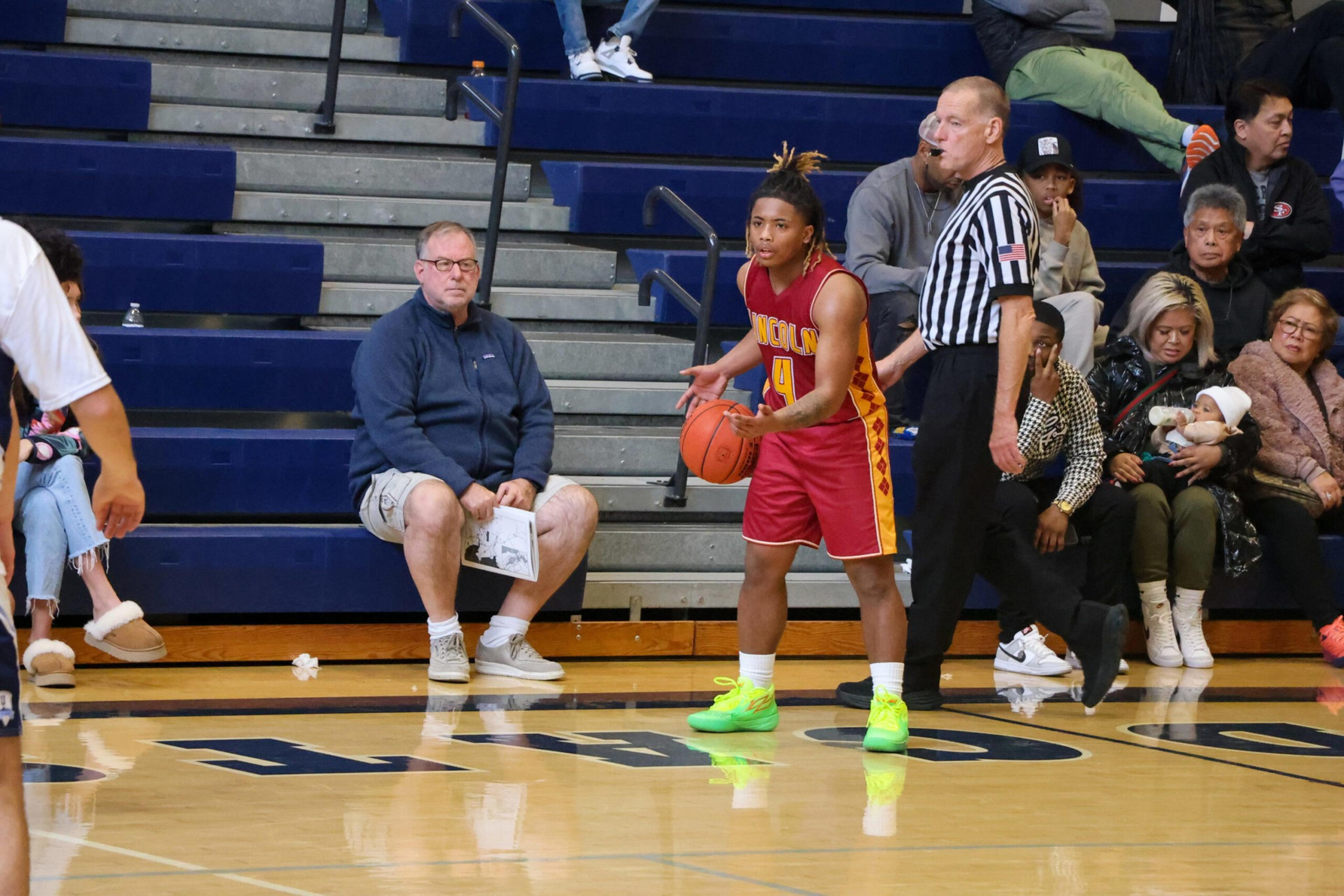 A basketball player in red and yellow holds the ball near the sideline while a referee and spectators watch from the bleachers.