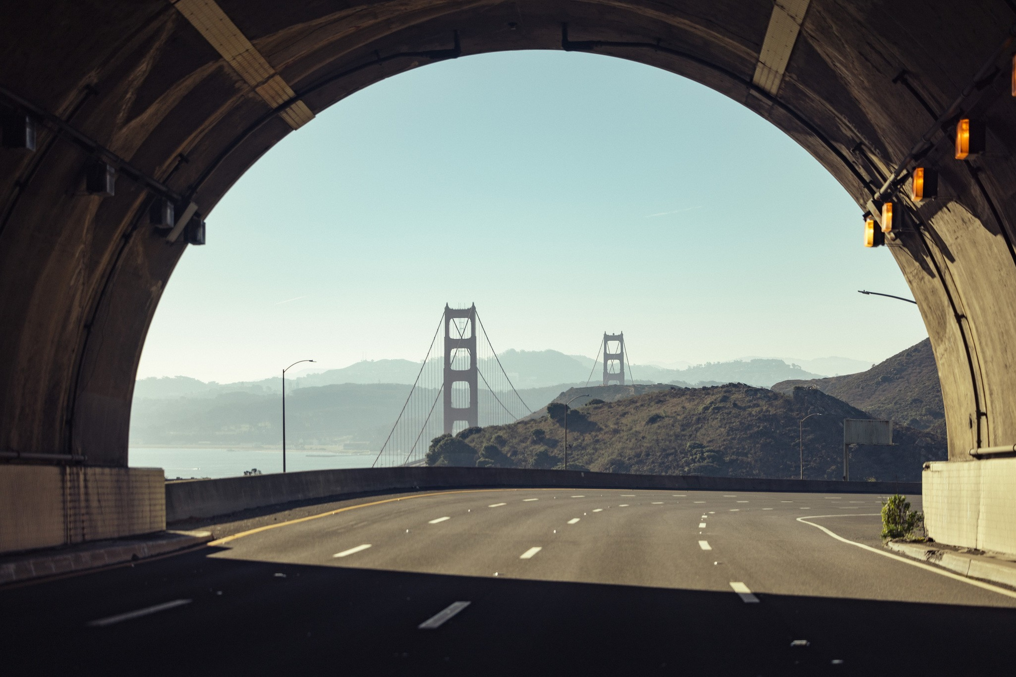 A clear highway curves out of a tunnel toward the Golden Gate Bridge, with hills and a calm sky in the background.