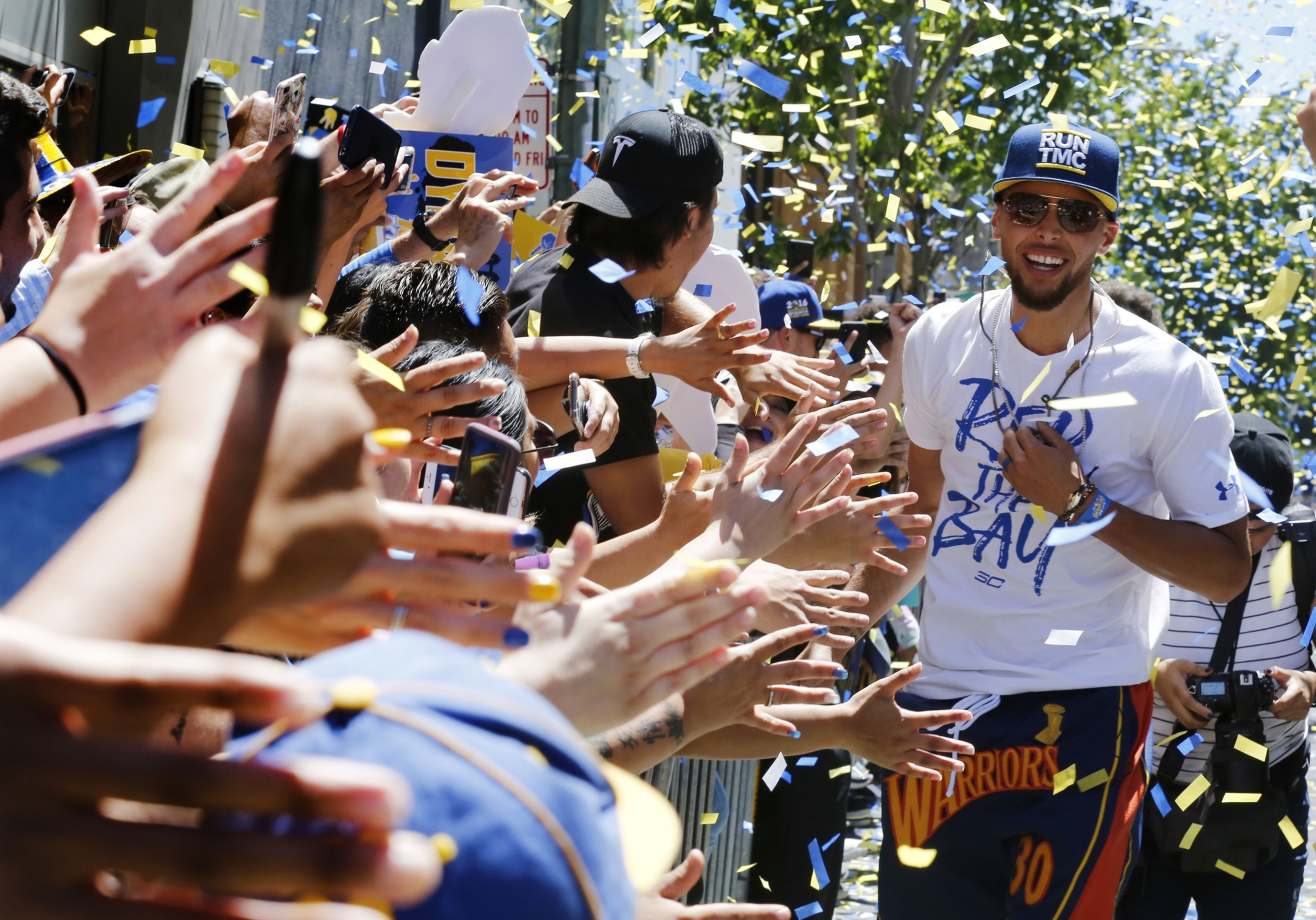A man in a white shirt and cap smiles, walking through a crowd with arms outstretched, surrounded by cheering fans and falling confetti.