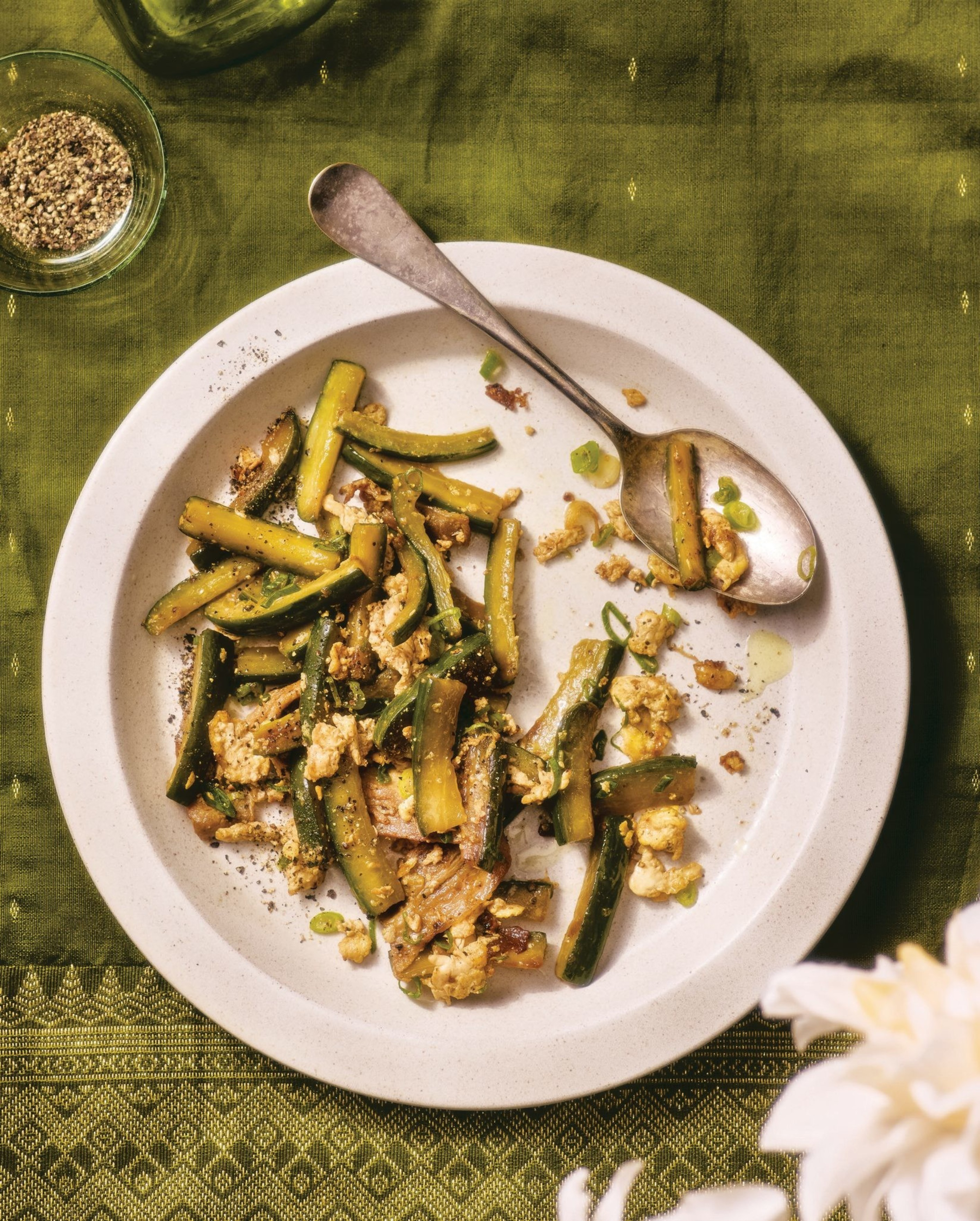 Stir-fried zucchini strips with scrambled egg pieces and green onion slices are on a white plate with a spoon, set on a green patterned cloth.