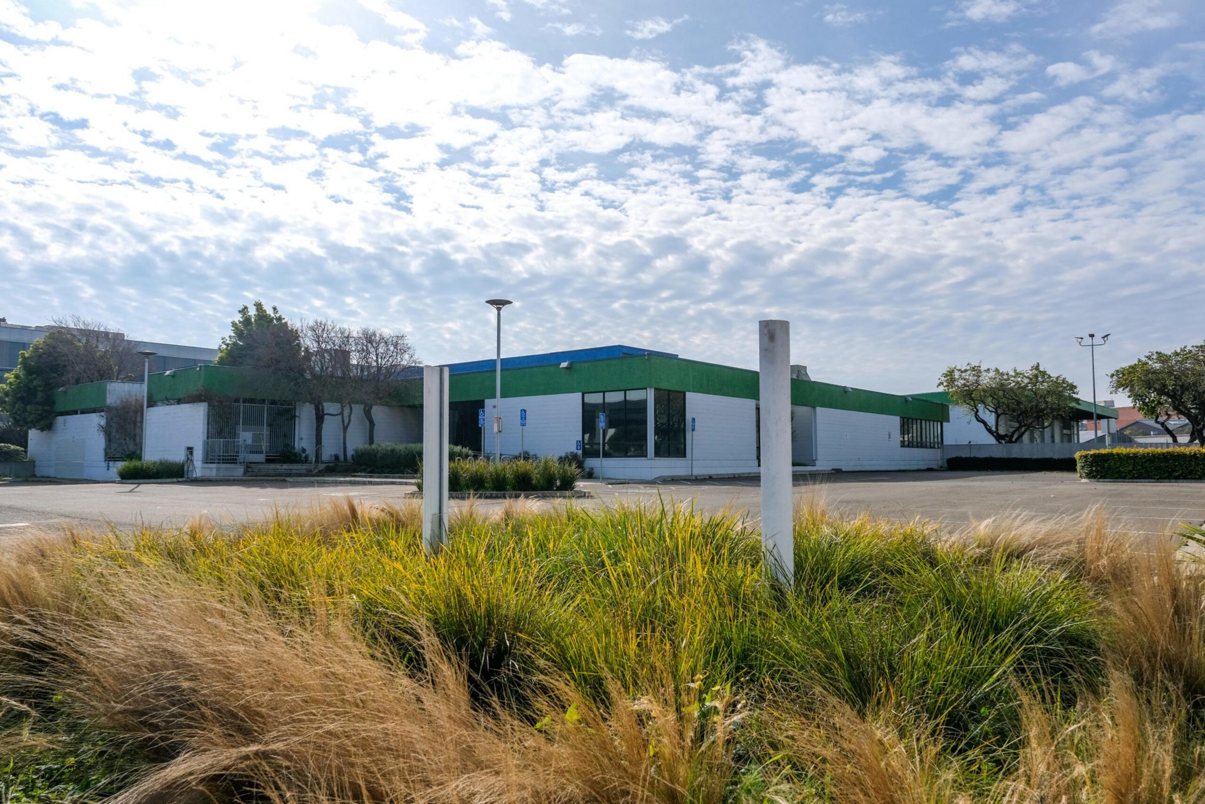 A single-story building with a flat roof and green trim is surrounded by an empty parking lot and ornamental grasses under a partly cloudy sky.