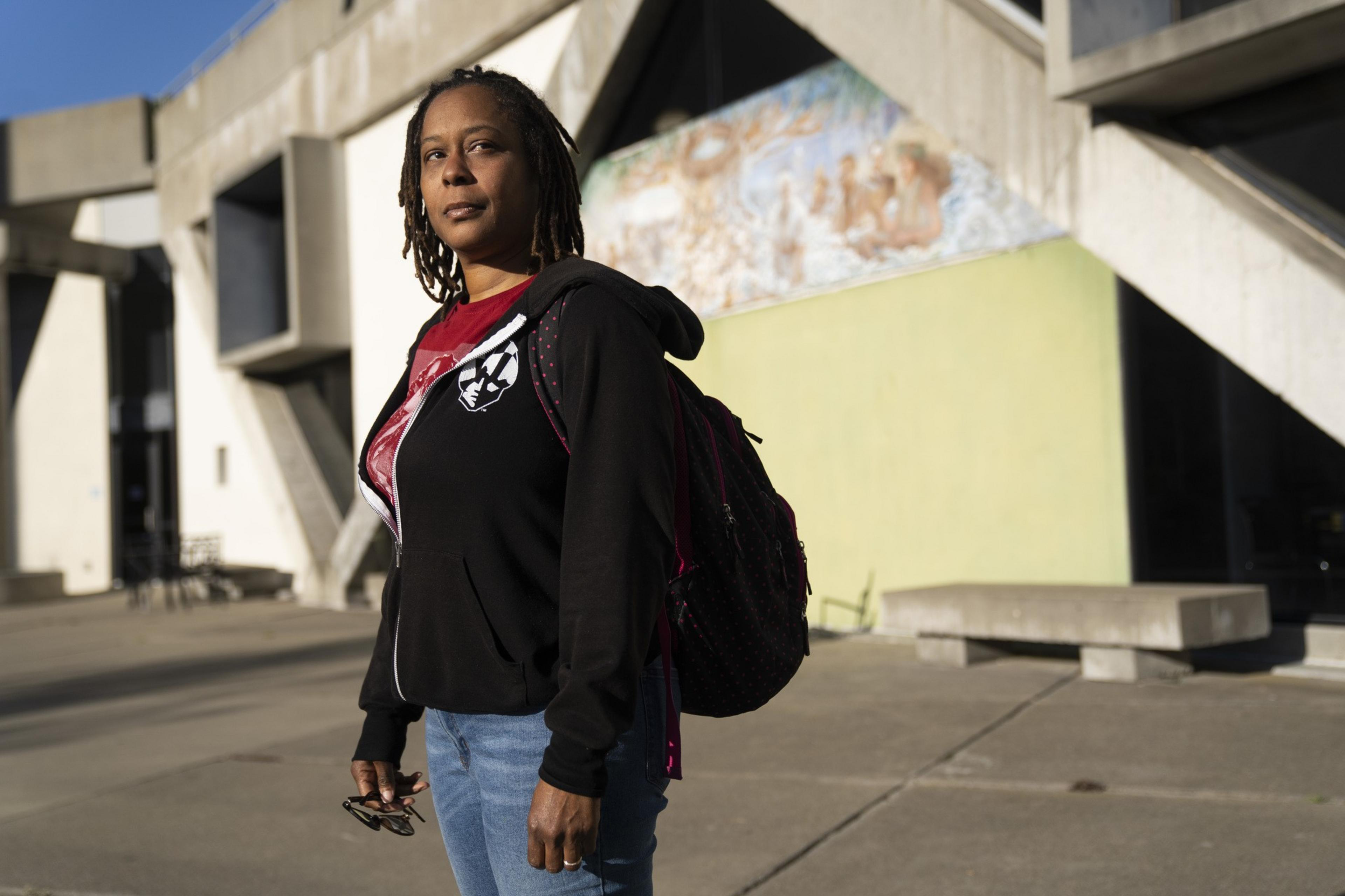 A person with dreadlocks, wearing a black hoodie and blue jeans, stands outside a building with geometric concrete architecture, under sunlight.
