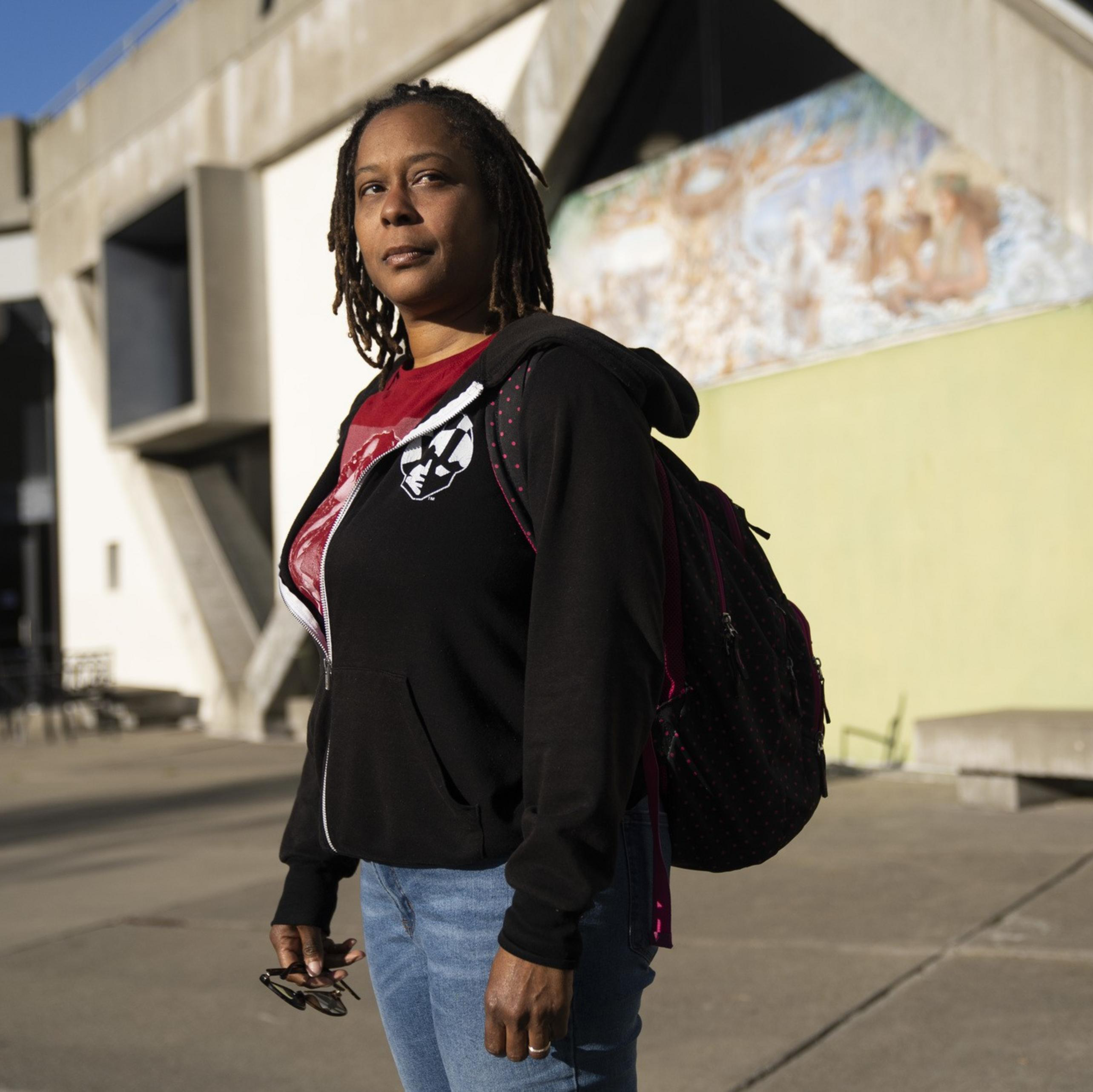 A person with dreadlocks, wearing a black hoodie and blue jeans, stands outside a building with geometric concrete architecture, under sunlight.