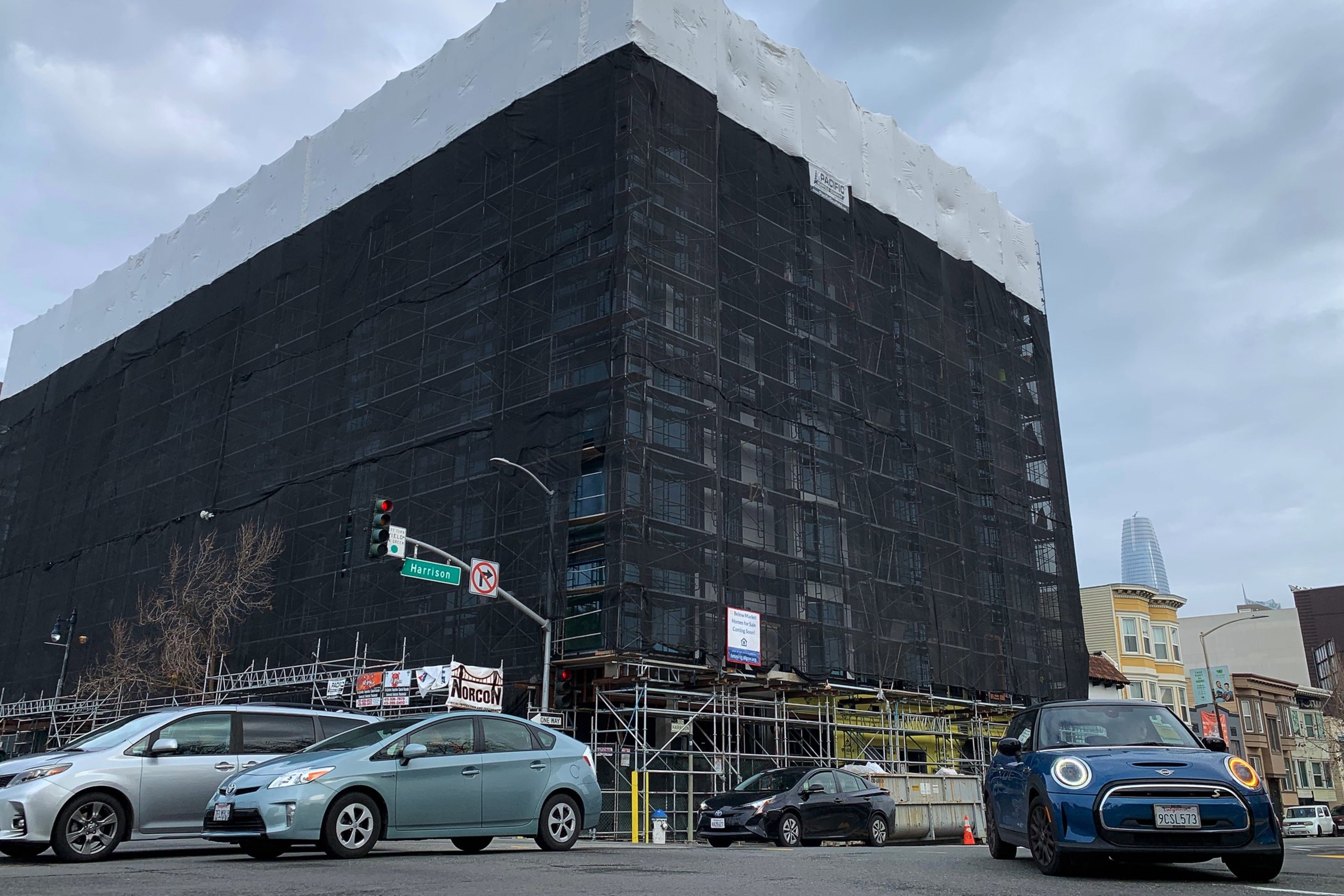 A large building under construction, covered with black scaffolding, with vehicles in the foreground.