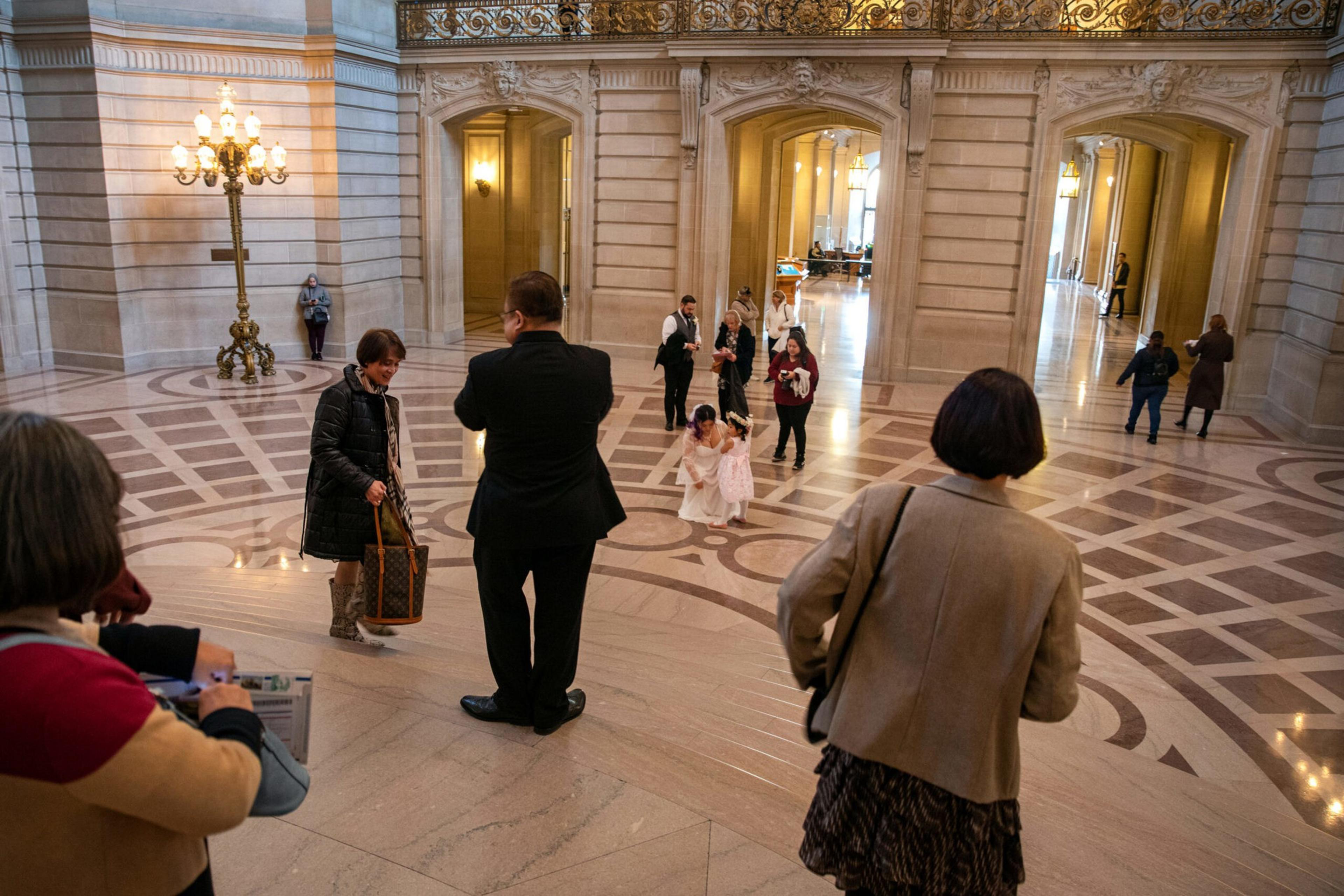 People are scattered in an ornate hall with marble floors and arches, including two little girls in white dresses playing near the center.