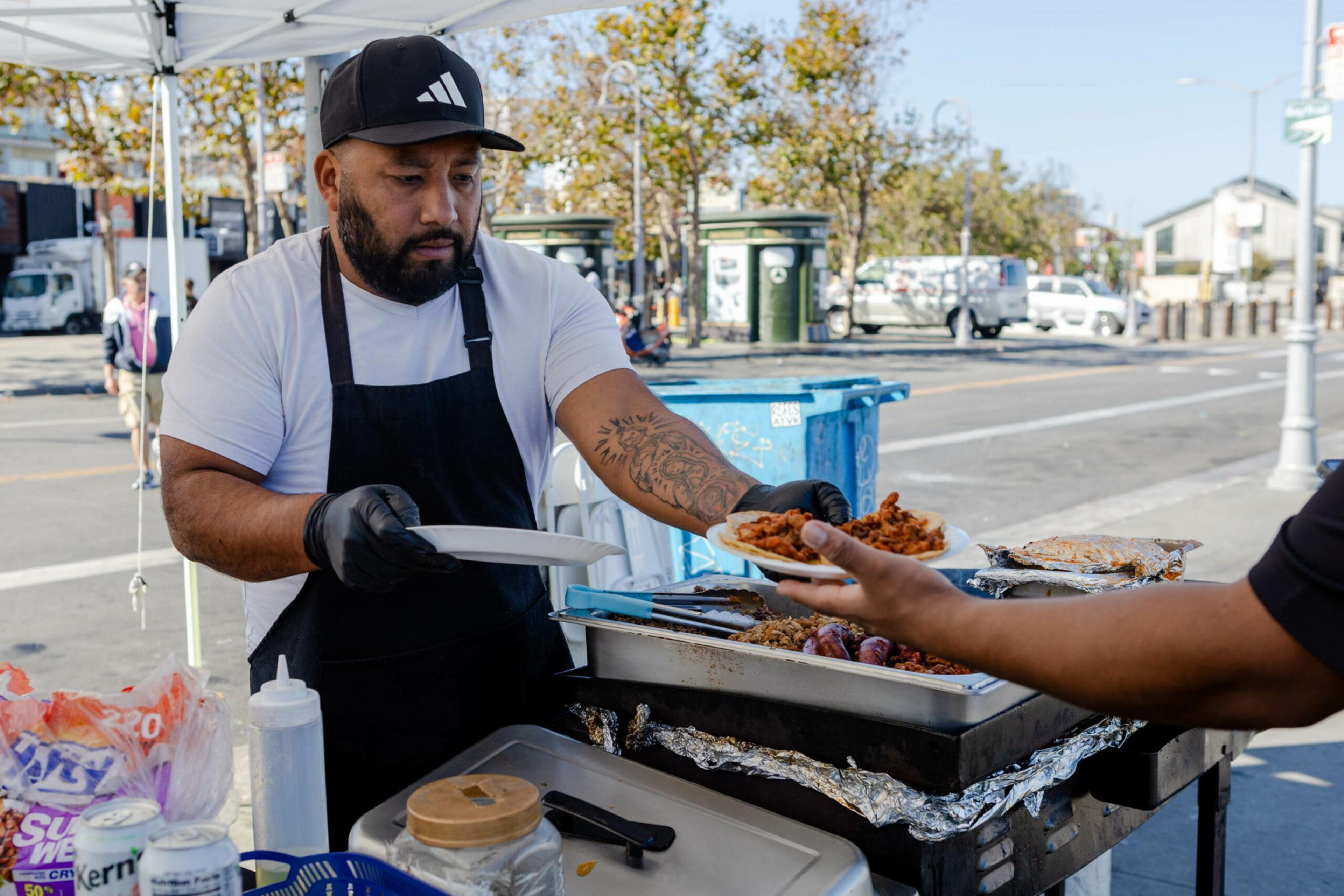 A bearded man in a black apron and cap serves food from a street food cart to a customer’s outstretched hand on a sunny day.